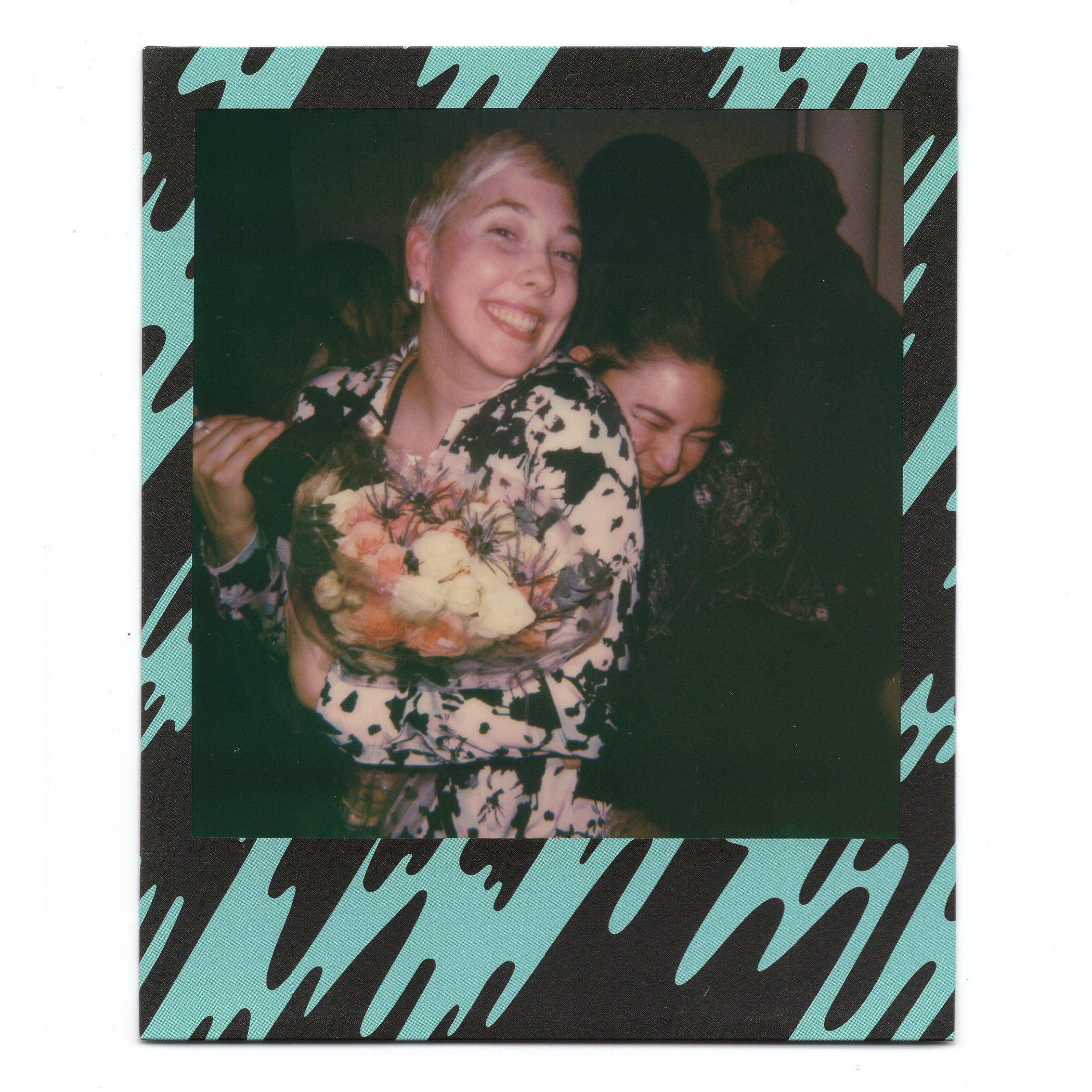 A woman smiling and holding a bouquet of flowers, with another woman hugging her from the side at a social event or party.