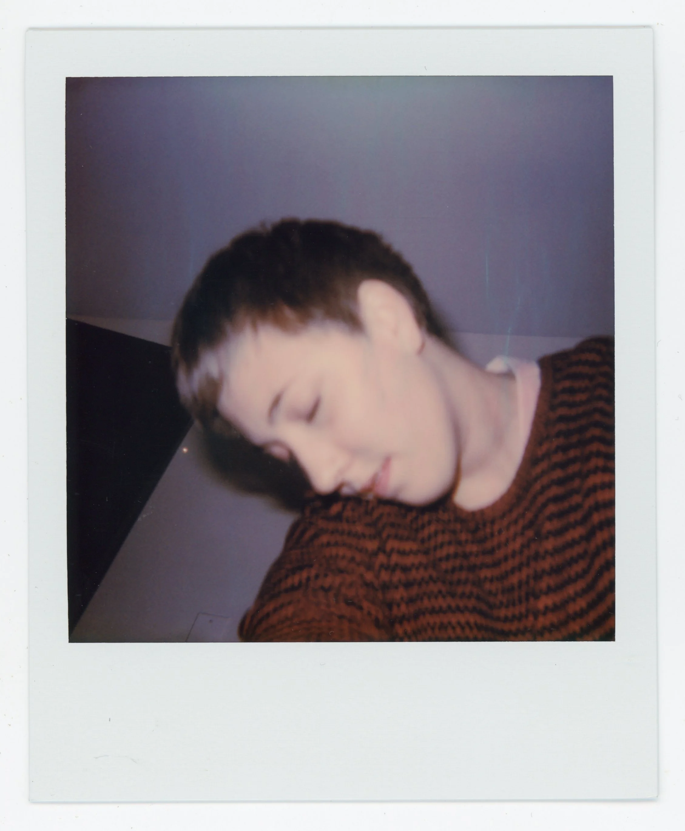 A young person with short dark hair is tilting their head with eyes closed, wearing a red and black striped sweater.