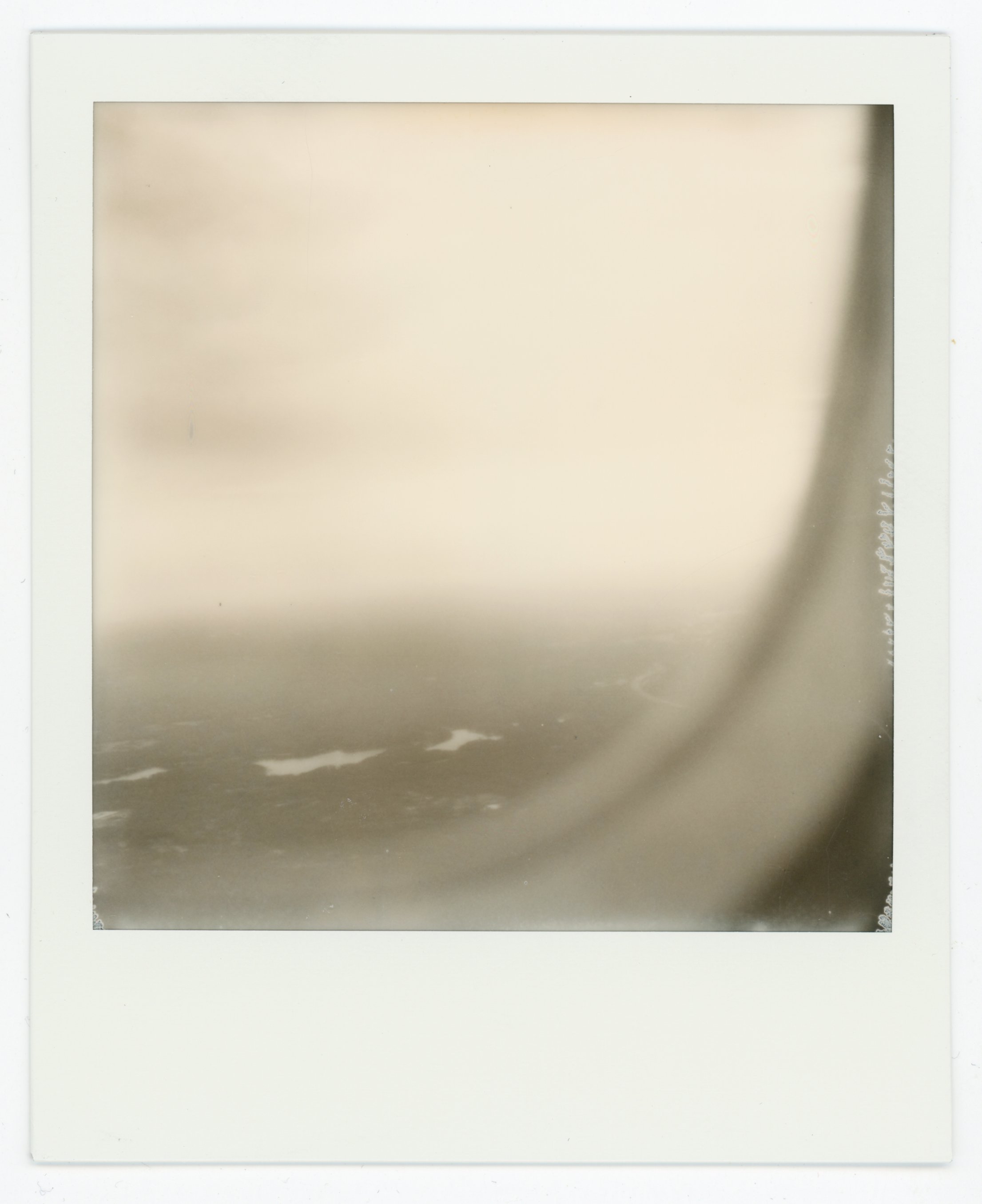 A Polaroid photo with a blurred sepia tone view of water and sky, with some waves visible at the bottom.