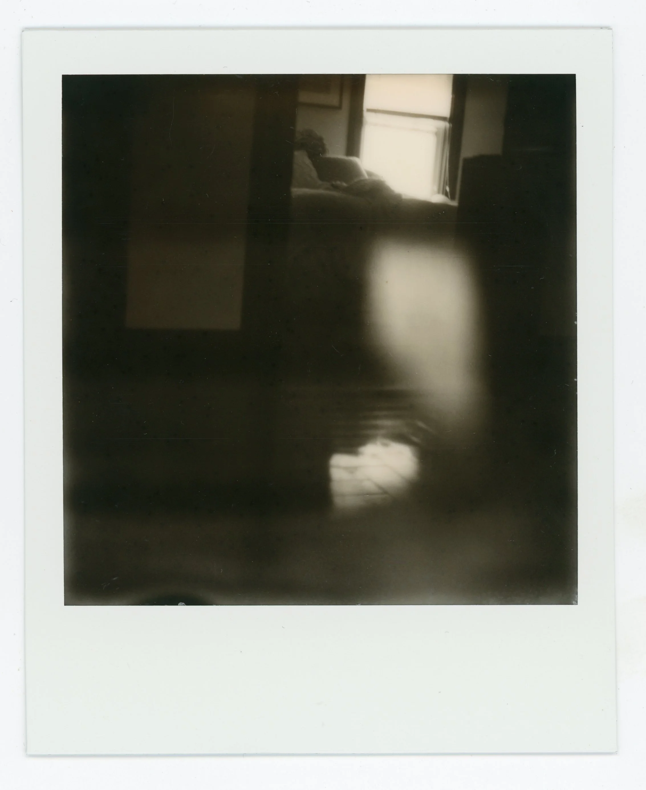 Blurred black-and-white photo of a person lying on a bed illuminated by daylight from a window, with a reflective floor in the foreground.