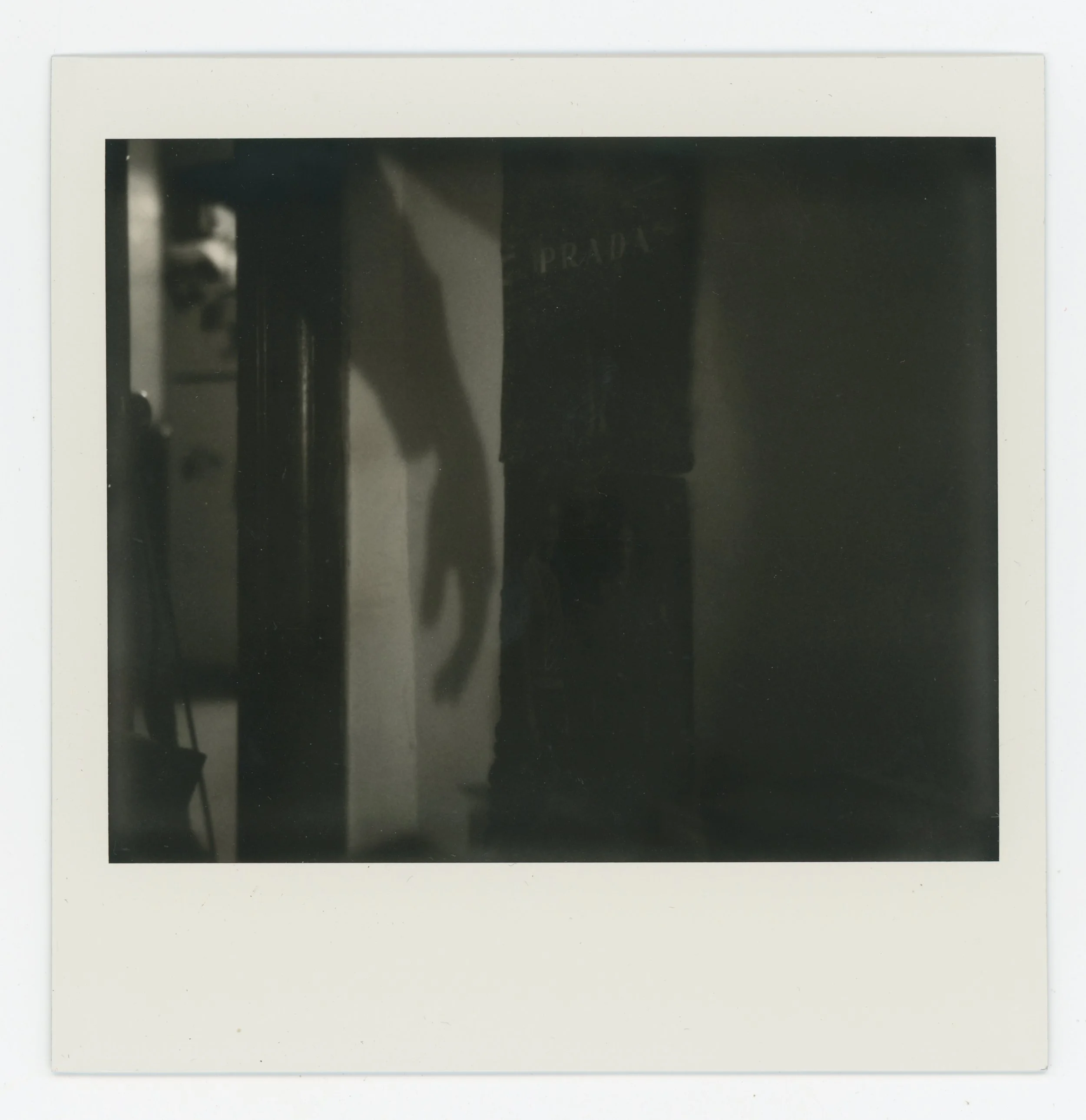 Black and white Polaroid photo showing a shadow of a hand on a wall, a partial view of a person in dark clothing with a Prada bag, and some furniture in the background.