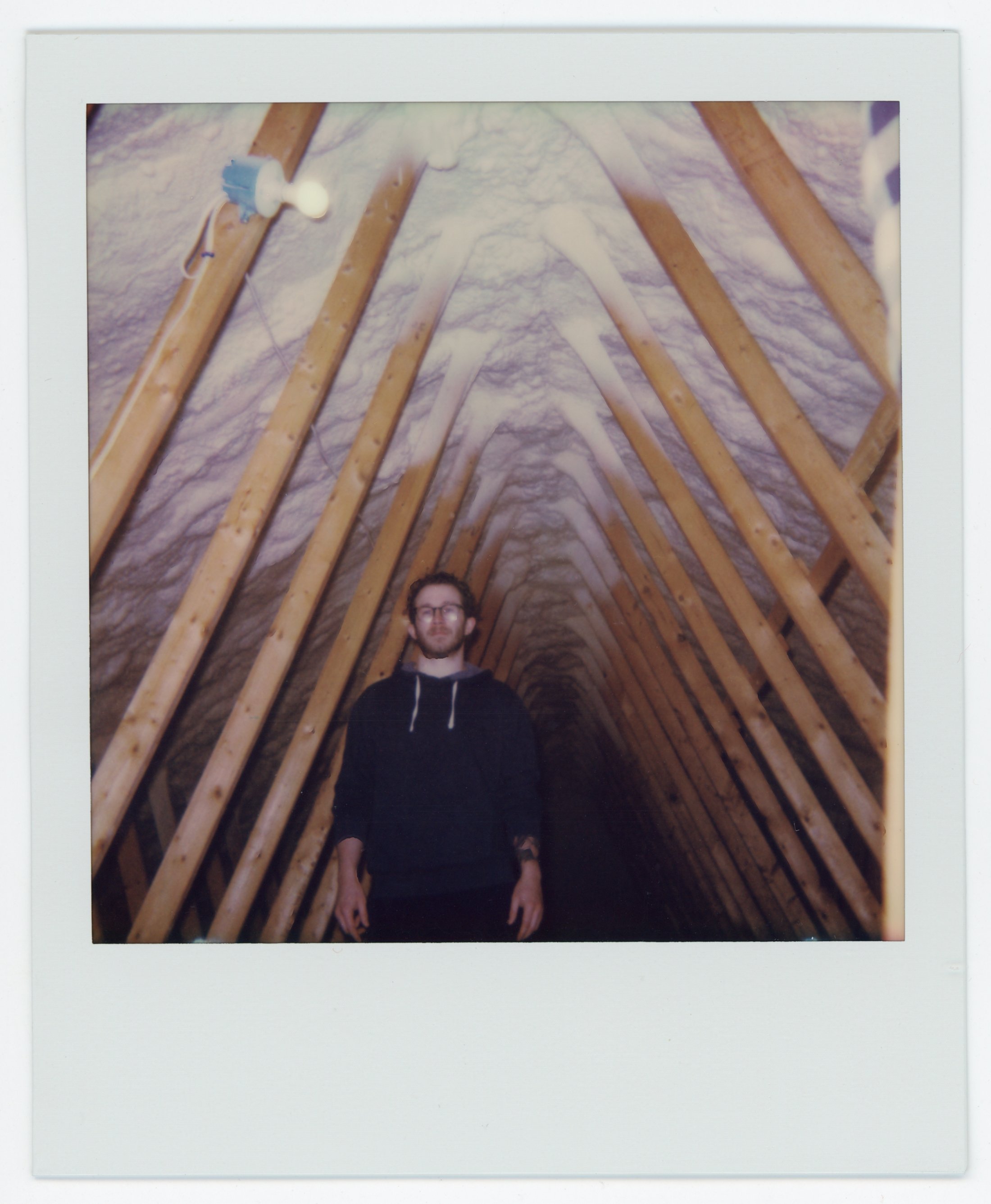 A man with glasses and a black hoodie standing in an attic with exposed wooden beams and foam insulation on the roof.