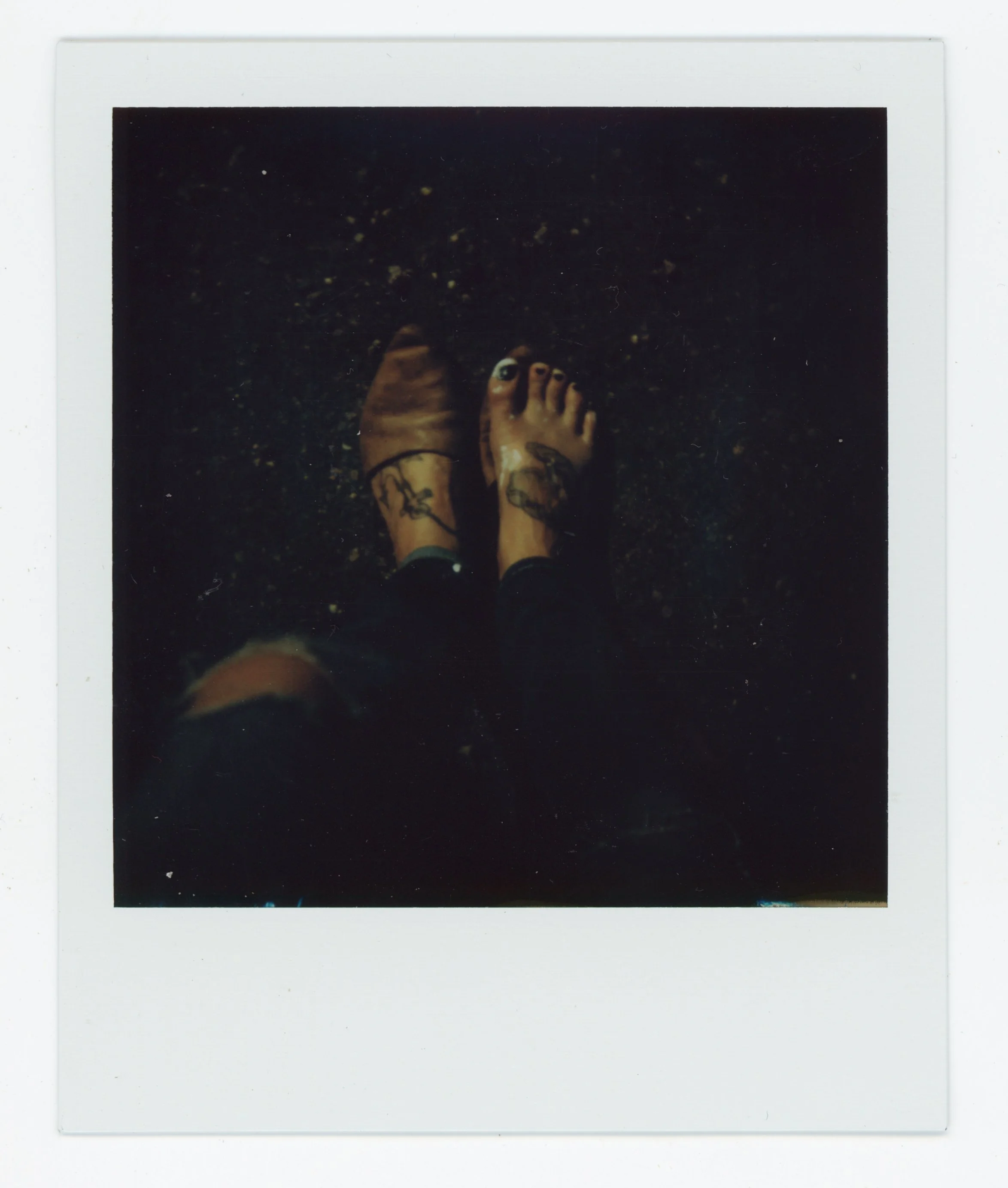 Polaroid photo showing a person's legs and feet, with visible tattoos, standing on a dark, textured surface.