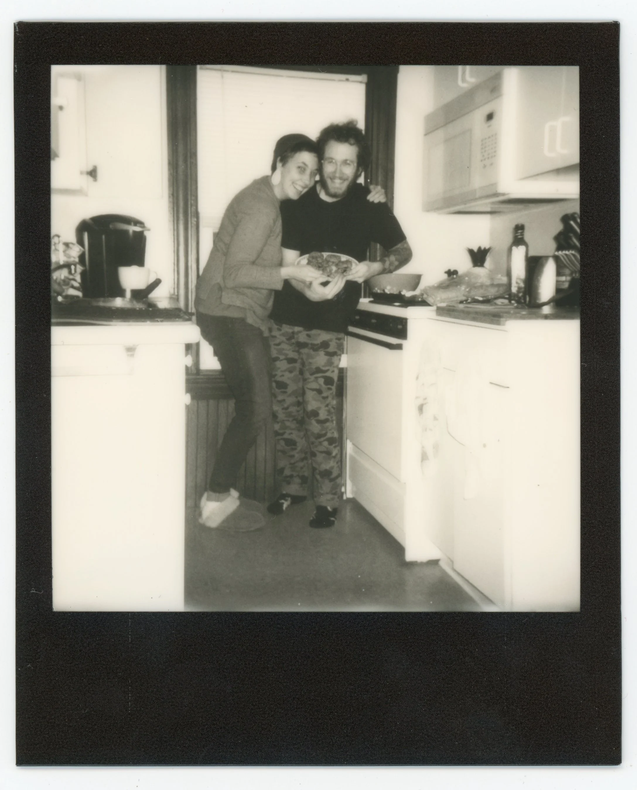 Two people standing in a kitchen, smiling and holding a plate of food, with various kitchen items visible on the counter.