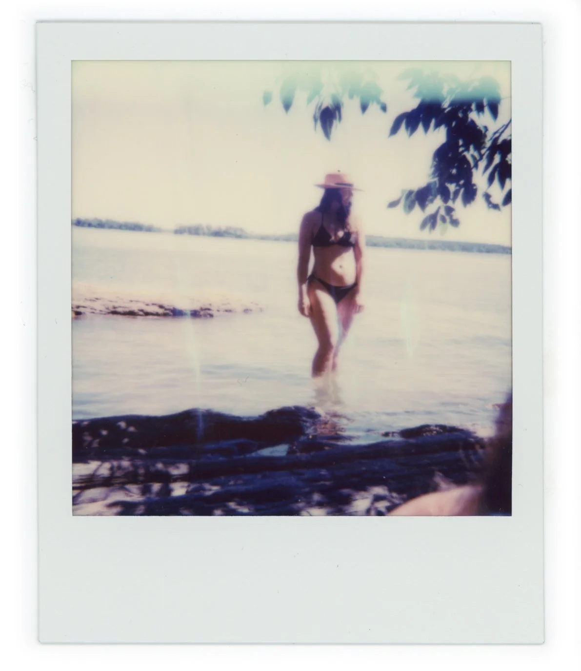 A woman in a bikini and sunhat standing in shallow water at the beach, with trees and a distant shoreline in the background.