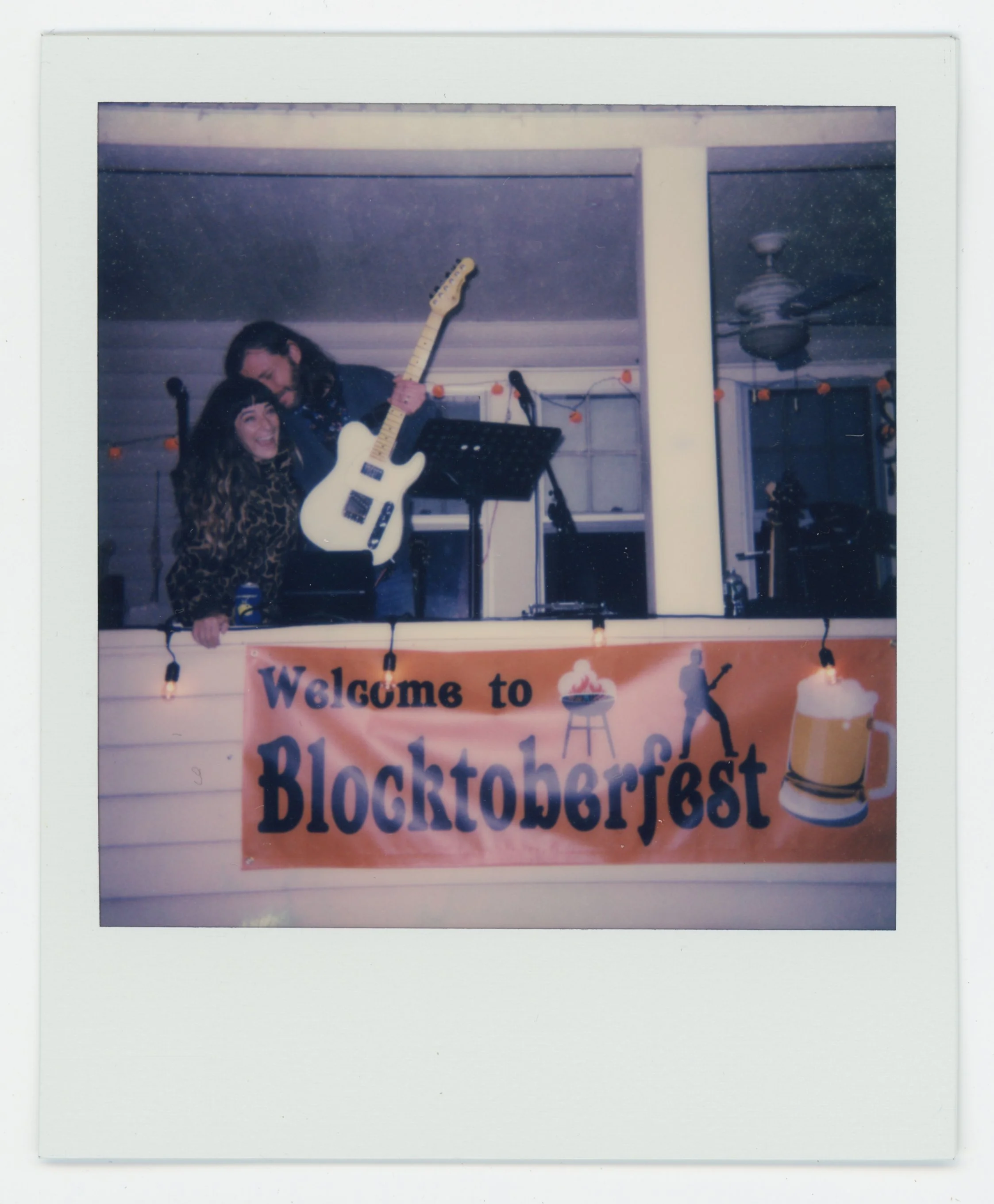 People on a porch with a banner reading 'Welcome to Blocktoberfest'. One person is holding a guitar. There are string lights and a ceiling fan in the background.