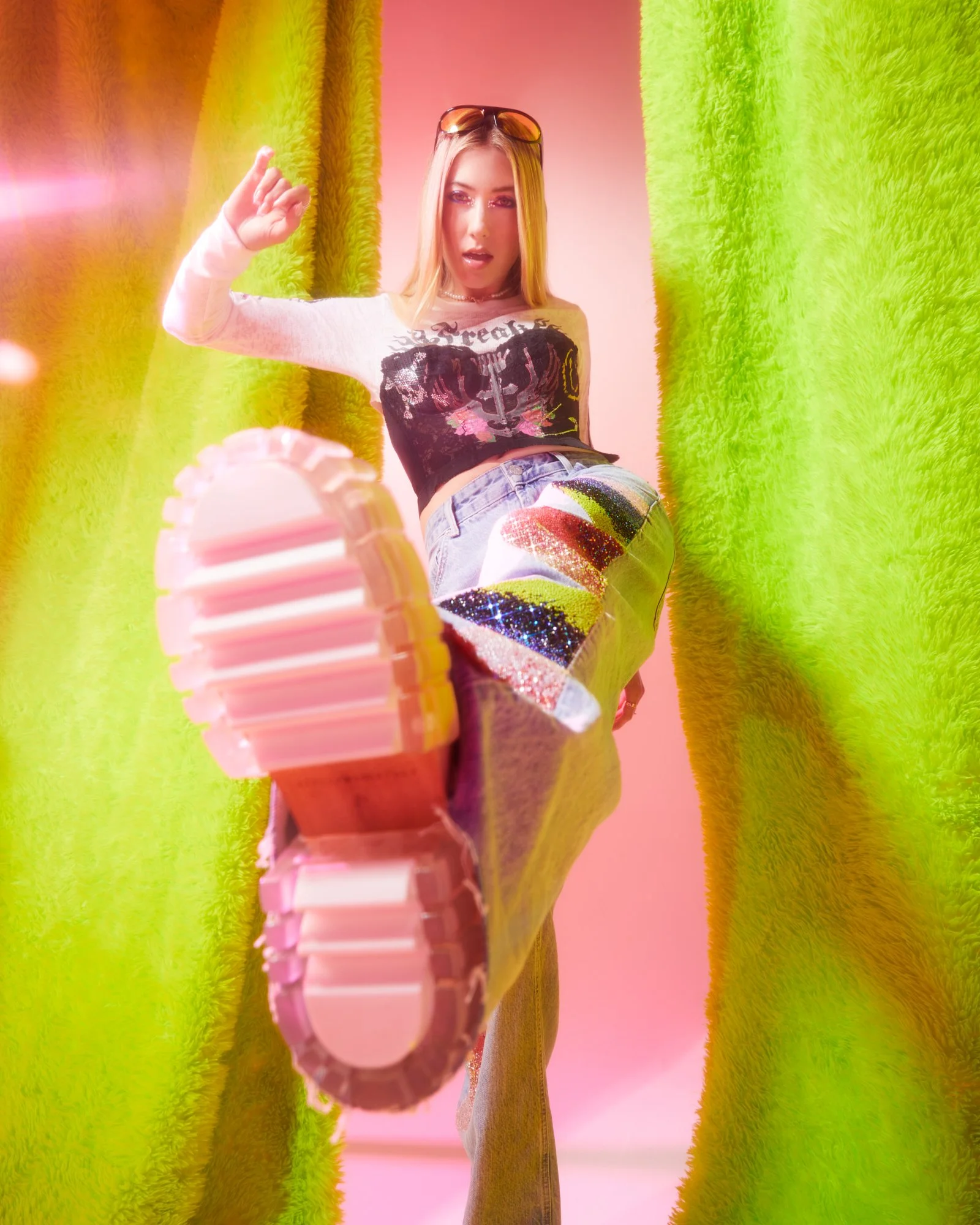 A young woman with blonde hair, wearing a black graphic crop top, colorful sequin patchwork jeans, sunglasses on her head, and a necklace, standing between green curtains, looking down at the camera while kicking her foot forward.