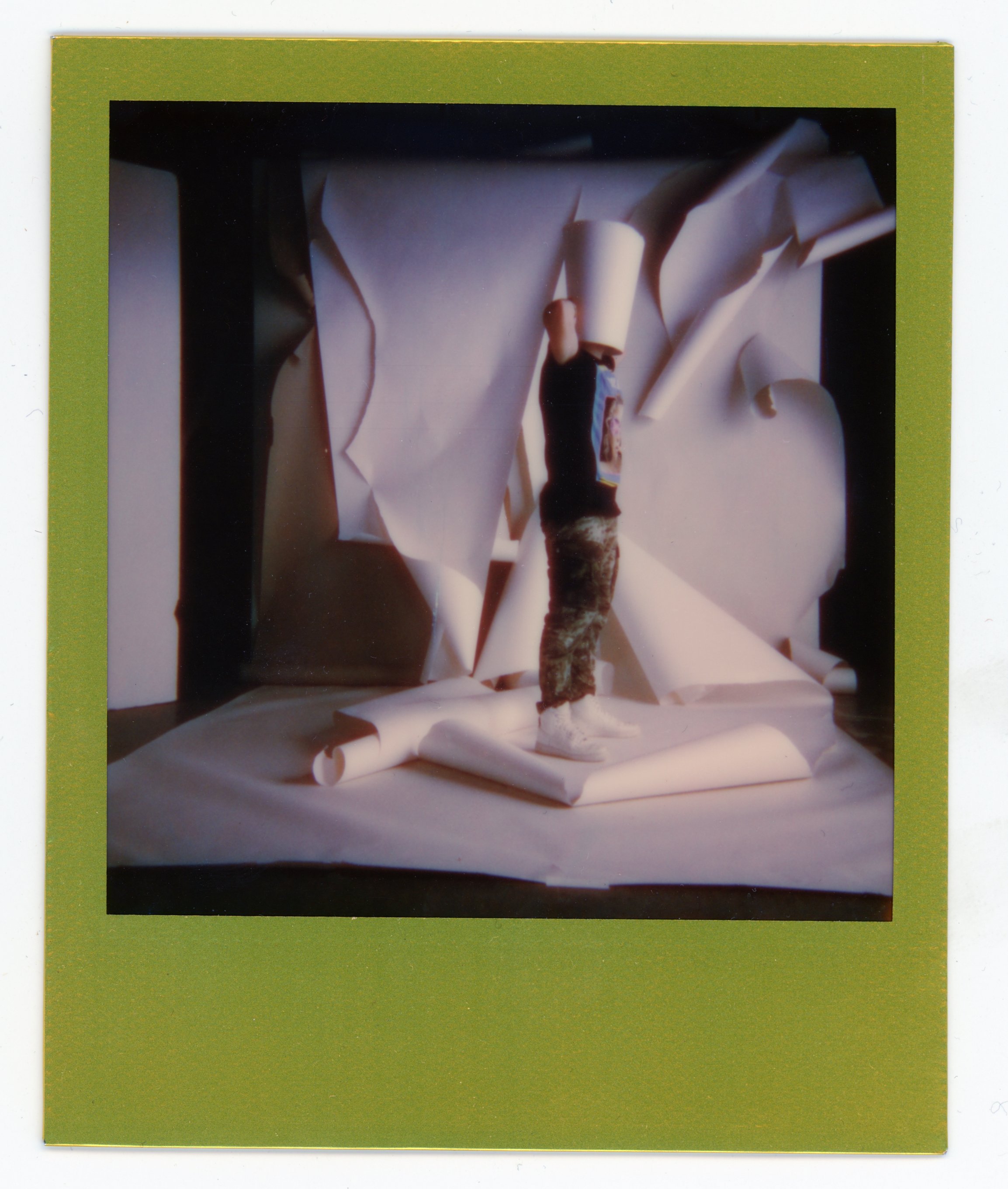 Abstract art piece with various rolled and crumpled papers, a small figure with a white hat, and a black figure with a camouflage pattern, all arranged on a white surface, bordered by a lime green frame.