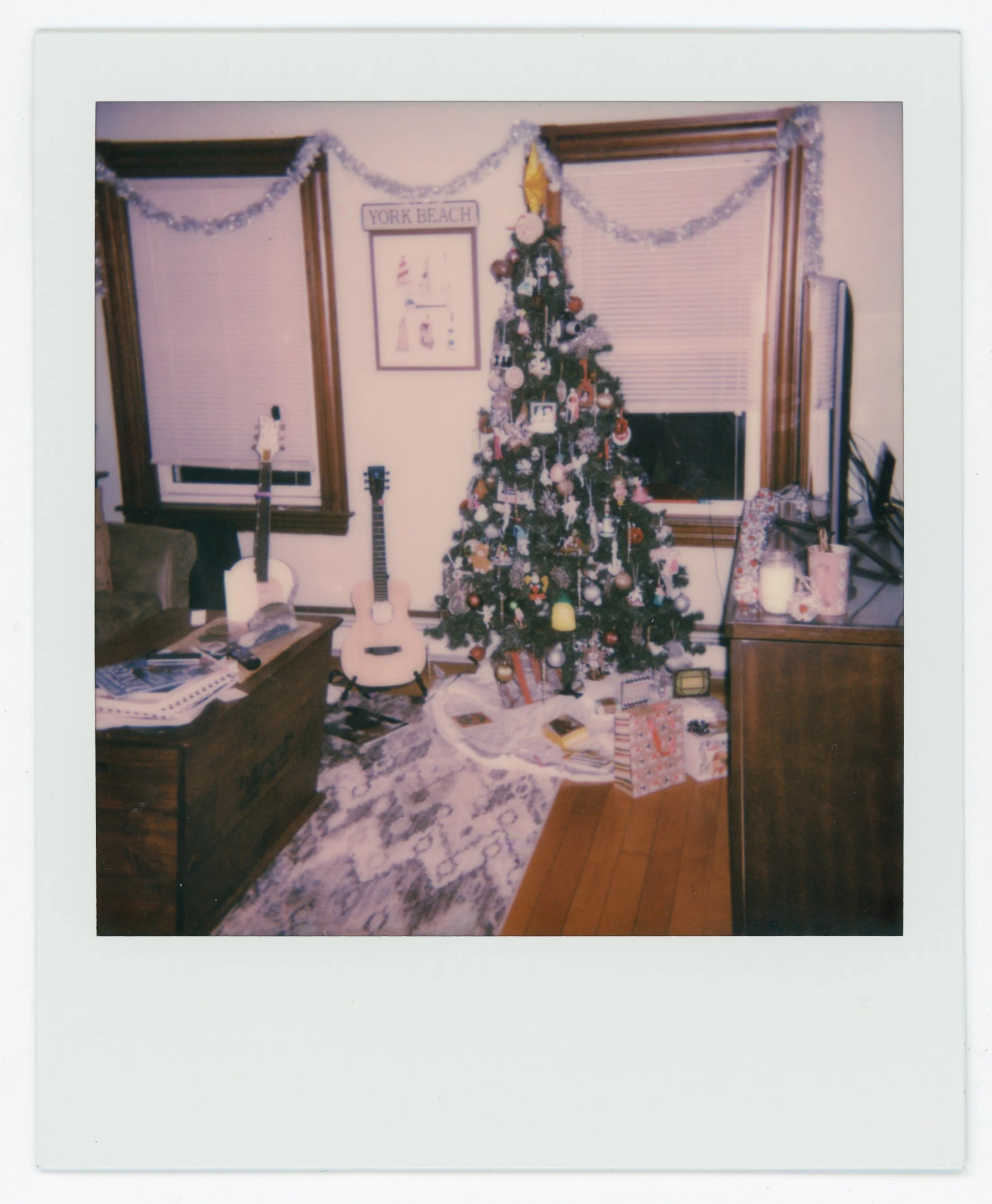 A decorated Christmas tree with presents underneath in a living room with two guitars near the window.