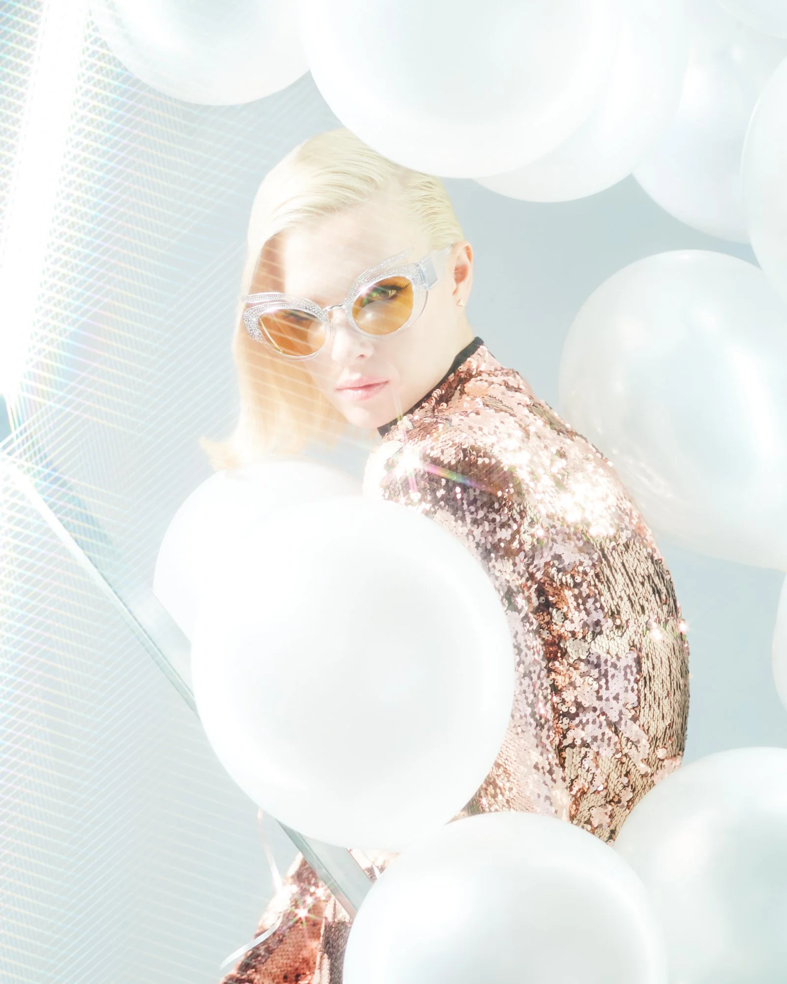 A woman with blonde hair wearing large, jeweled sunglasses and a sparkling, pink sequined jacket, surrounded by white balloons and reflective geometric patterns.