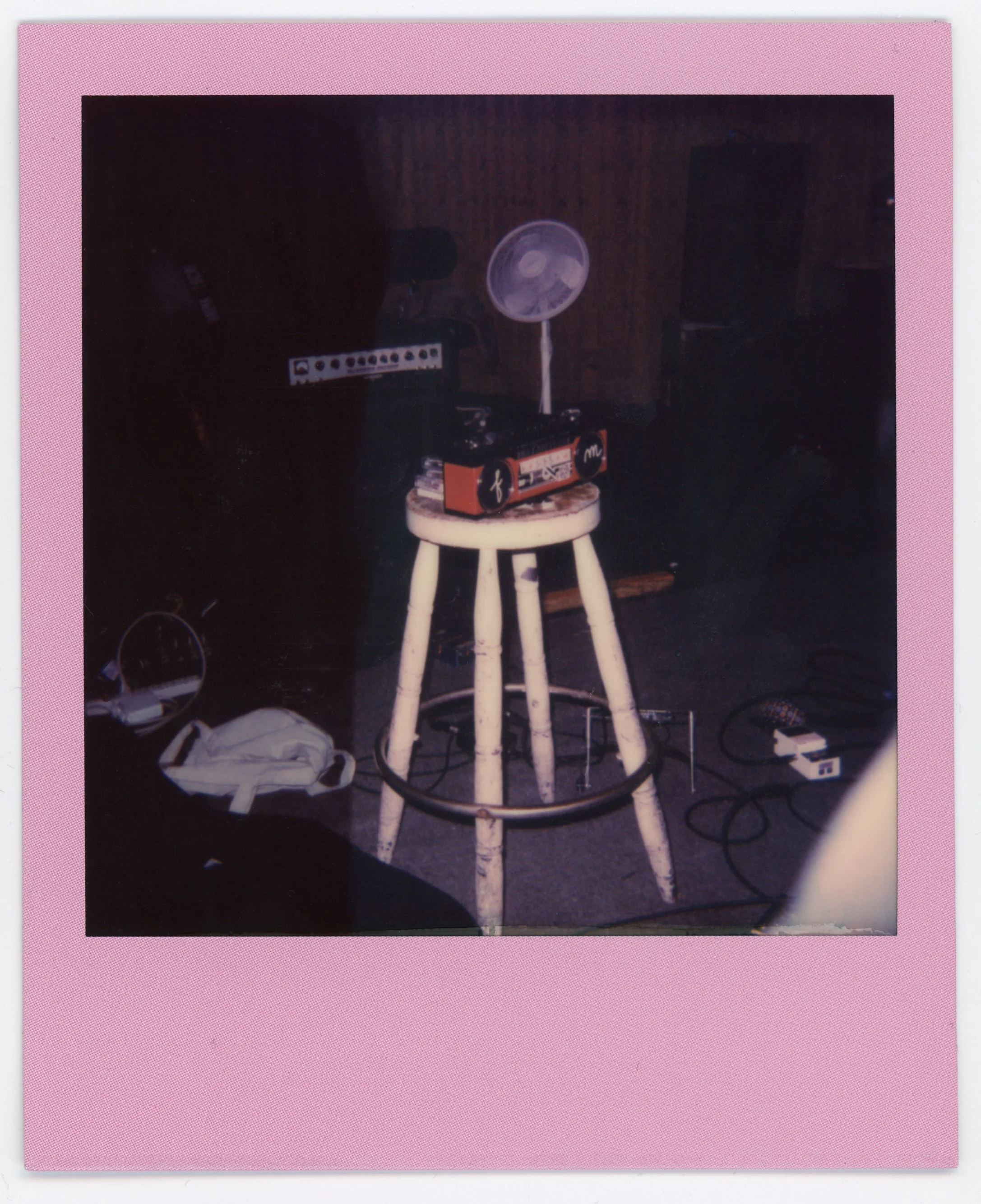 A white wooden stool with a vintage radio, a small electric fan, and some electronic equipment on top, in a dim room with musical equipment and wires on the floor.