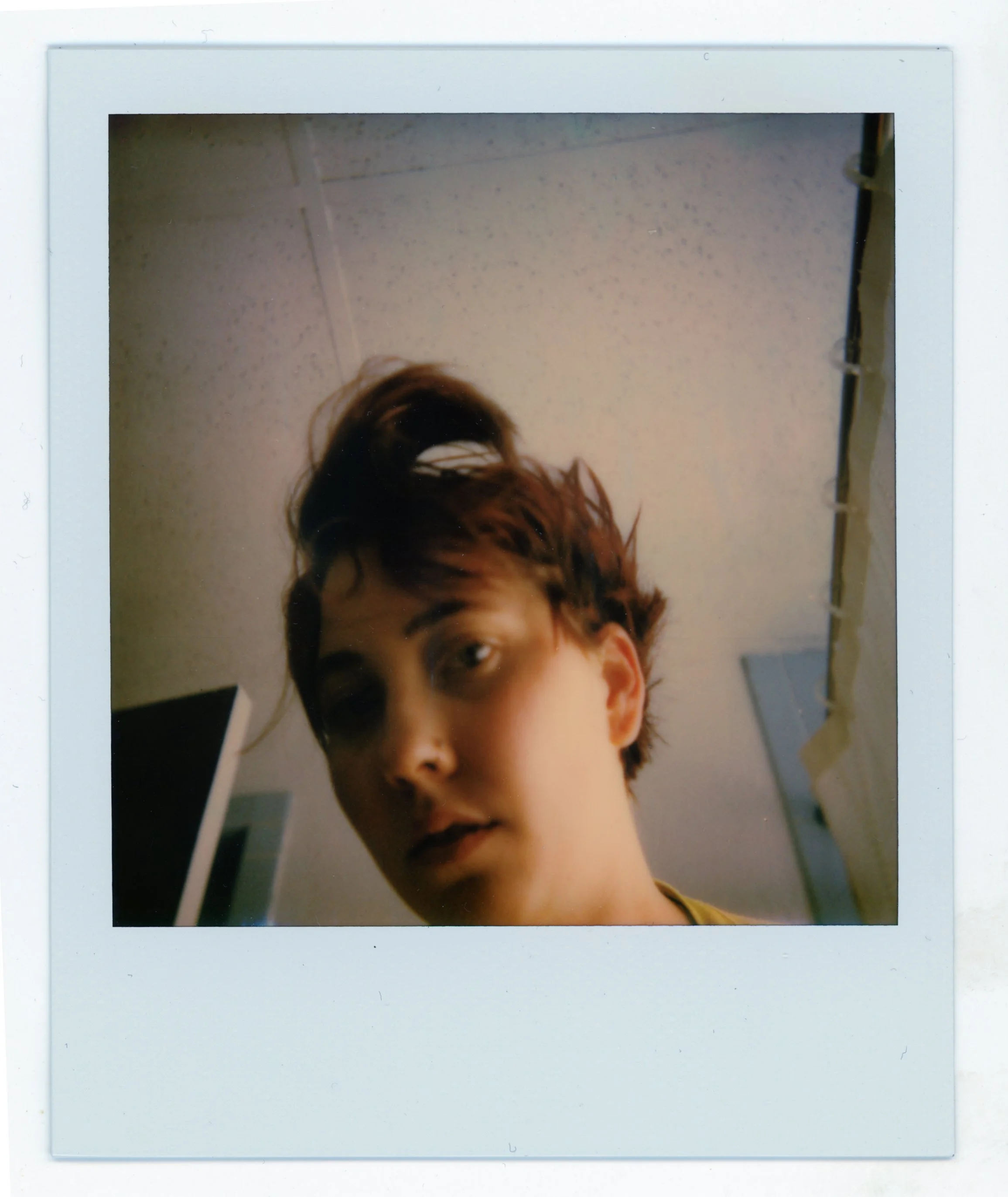 A close-up Polaroid photo of a young person with short, tousled hair, looking at the camera with a neutral expression, in a room with a textured ceiling and a dark doorway.