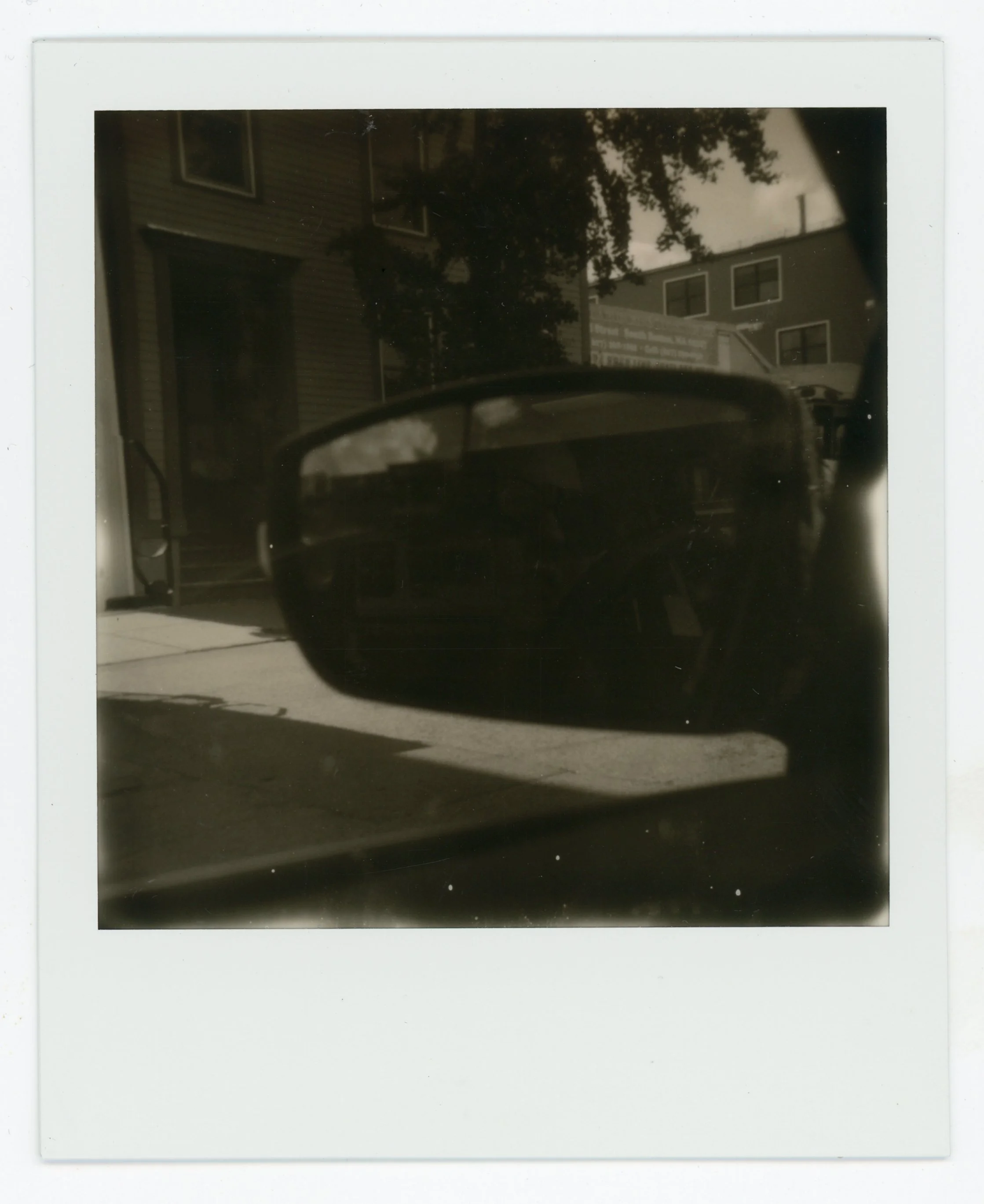Black and white photograph of a side view mirror of a car, with buildings, a sidewalk, and trees visible in the background.