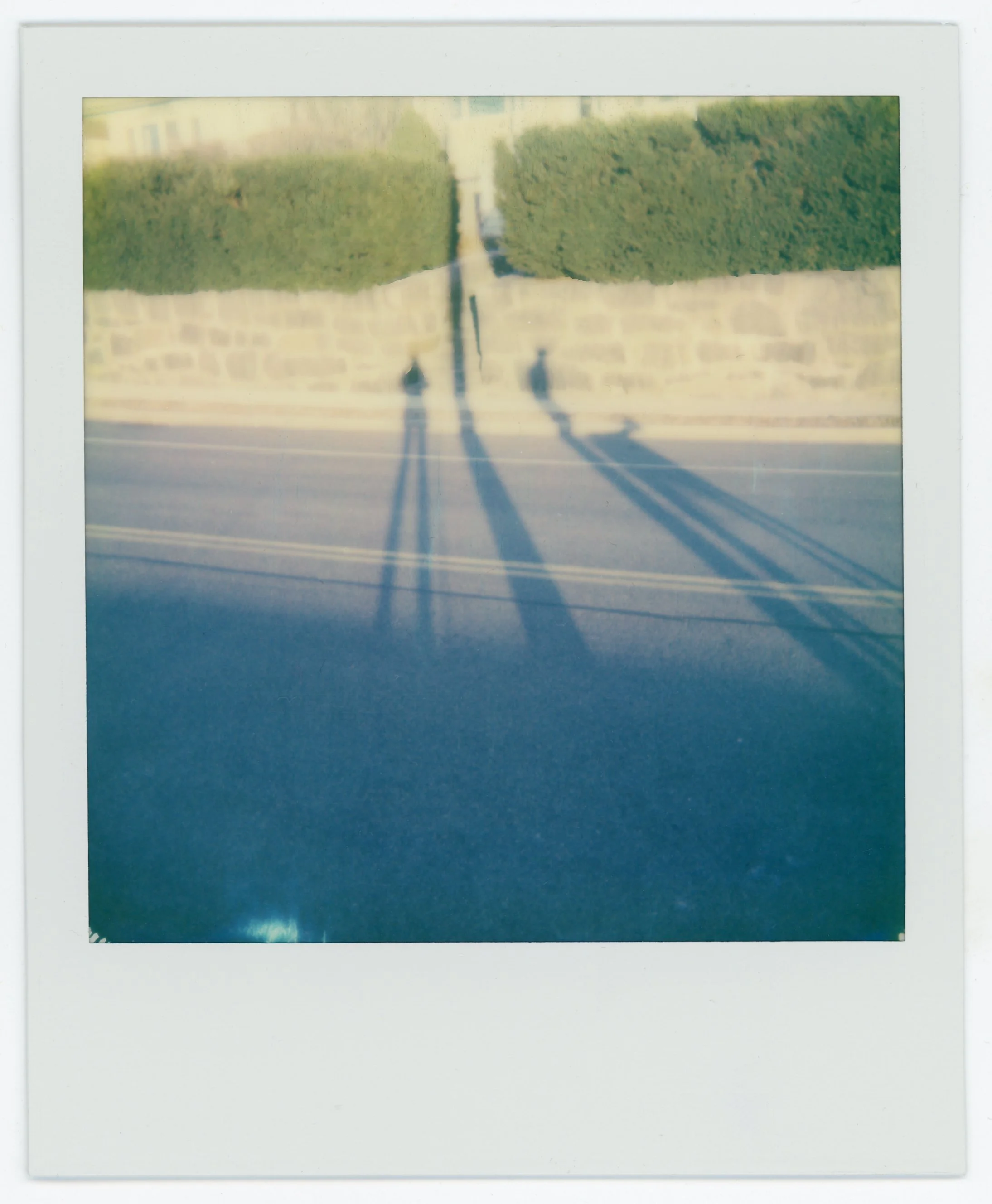 Polaroid photo showing the shadows of two people cast on the street with a stone wall and green bushes in the background.
