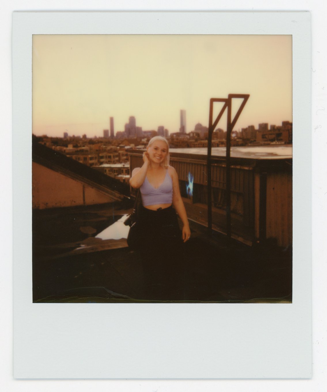 A woman smiling on a rooftop at sunset with a city skyline in the background.