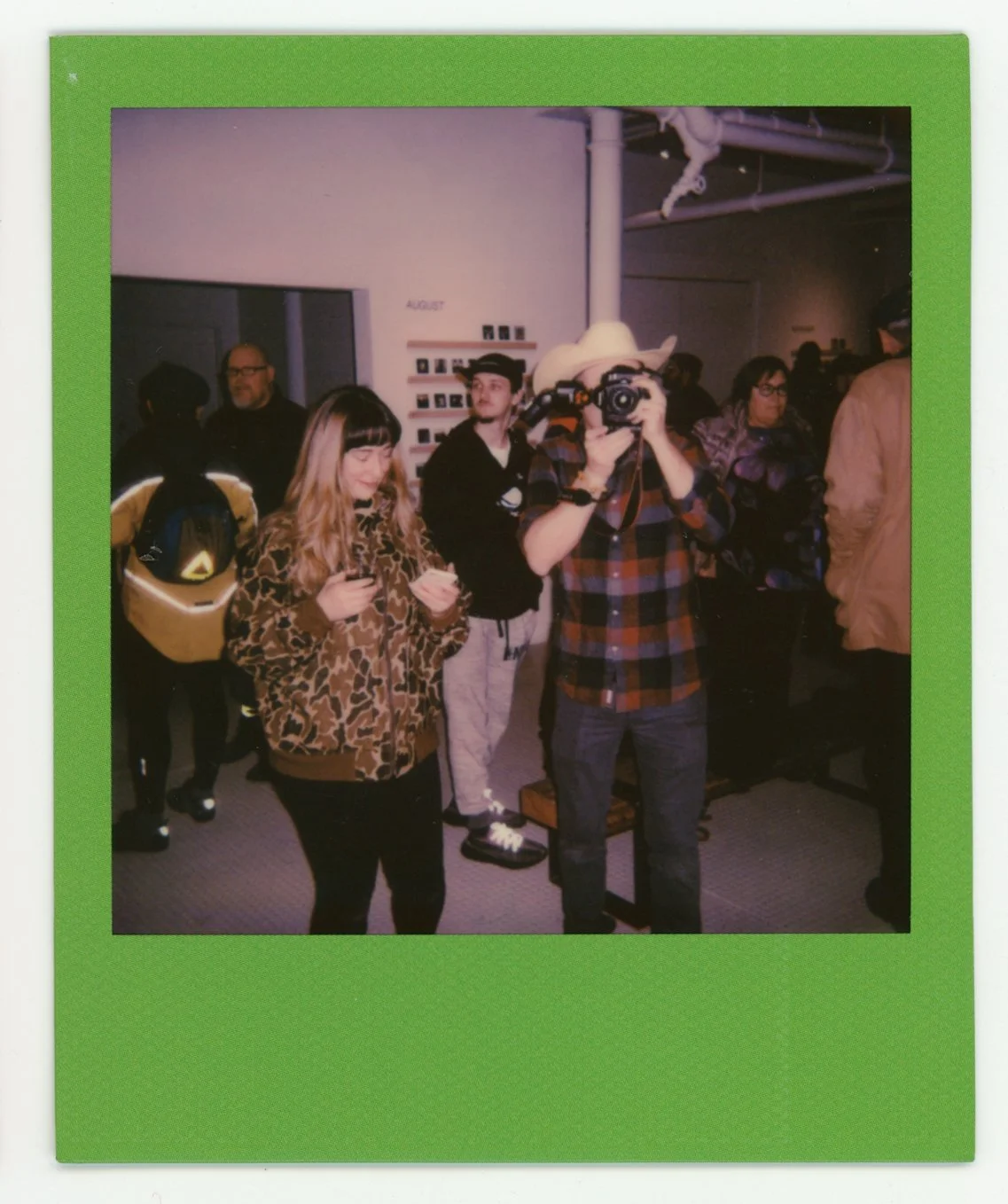 People at an indoor event, with one person taking a photograph and others standing around, some looking at the camera, in front of a white wall with a small display and the word 'AUGUST' written on it.