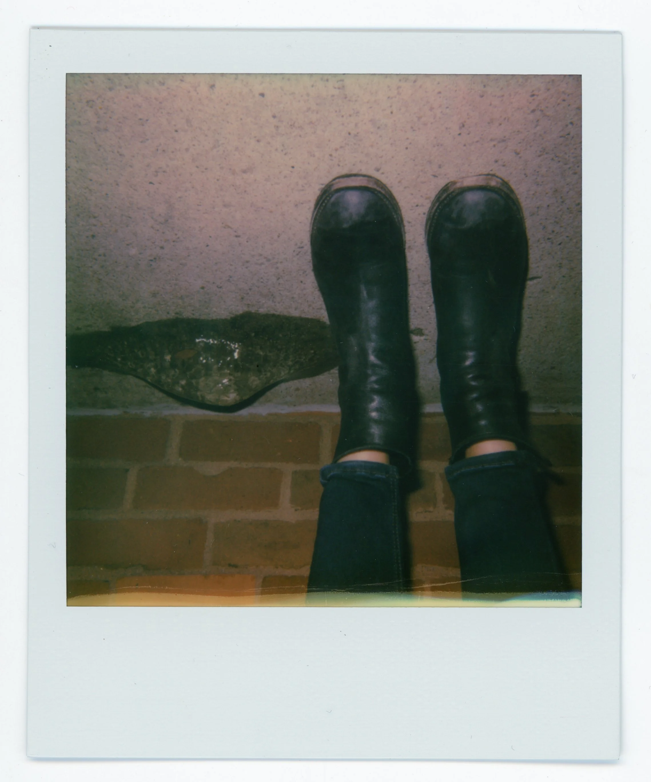 Person standing on a brick floor wearing black boots and jeans, with a stone and concrete wall in the background.