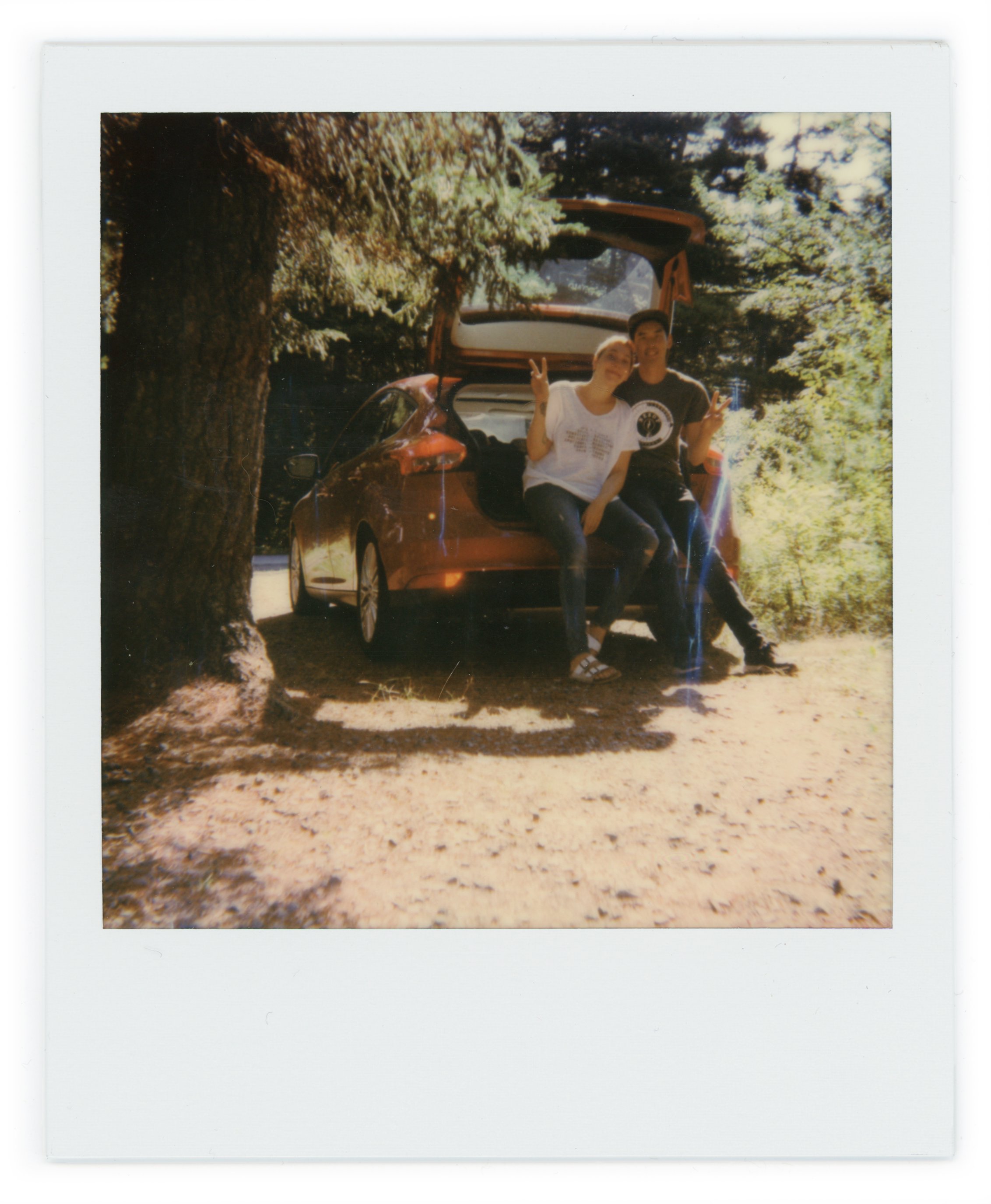 Two people sitting on the open trunk of a red car, making peace signs, in a wooded outdoor setting.