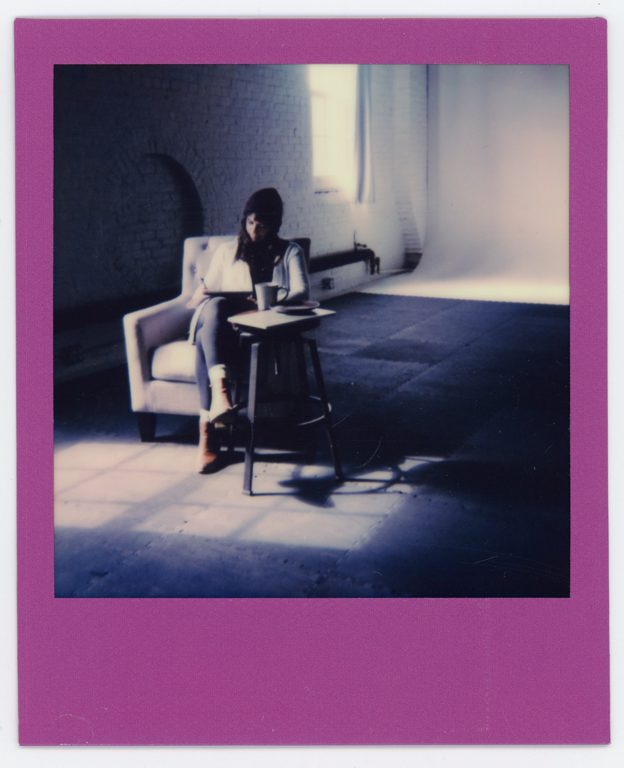 A woman sitting in a white armchair reading a book in a room with exposed brick walls, illuminated by natural light coming through a window.