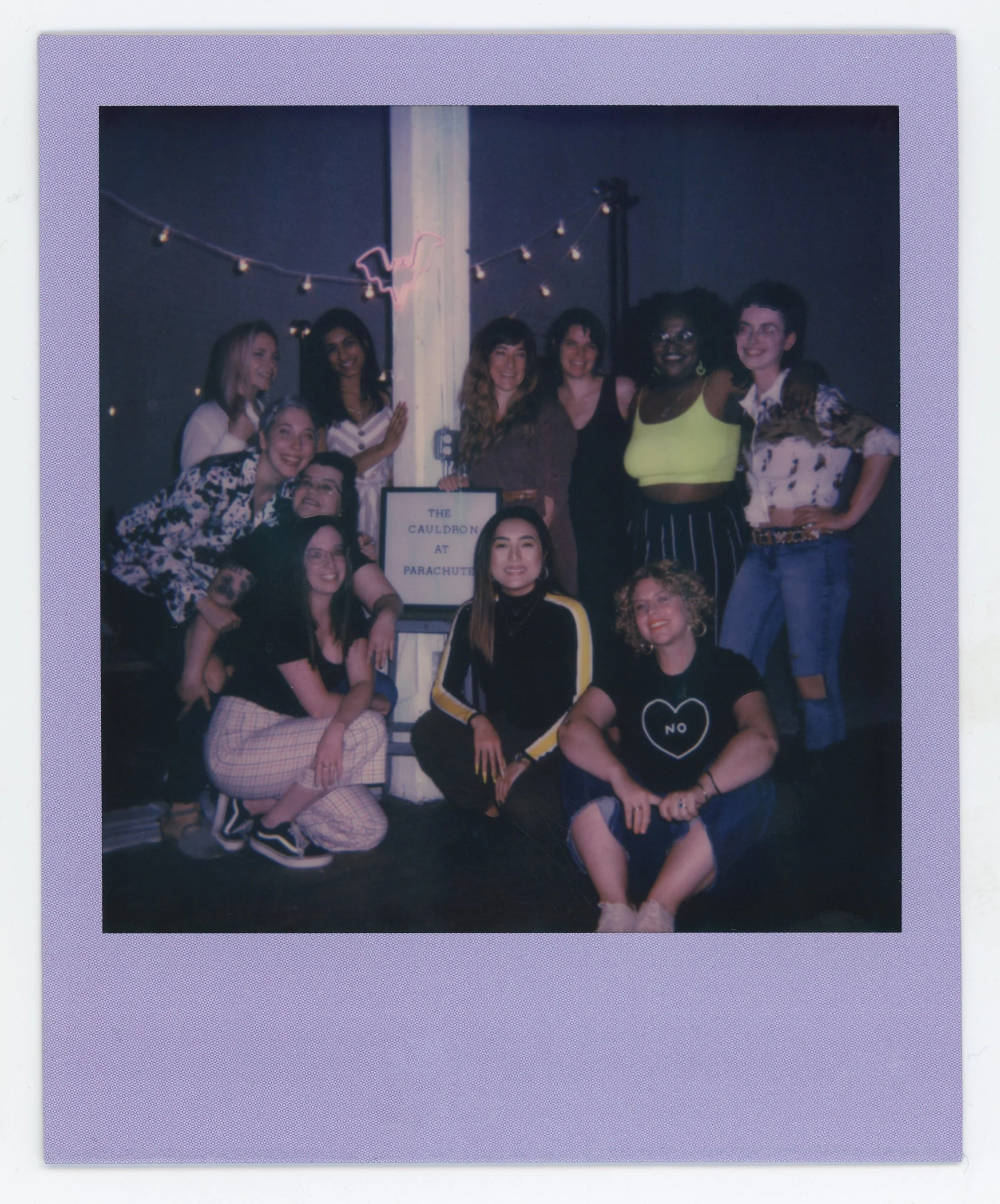 Group of women posing together at night in front of a sign that reads 'The Cauldron at Parachute,' with string lights and a neon flamingo in the background.