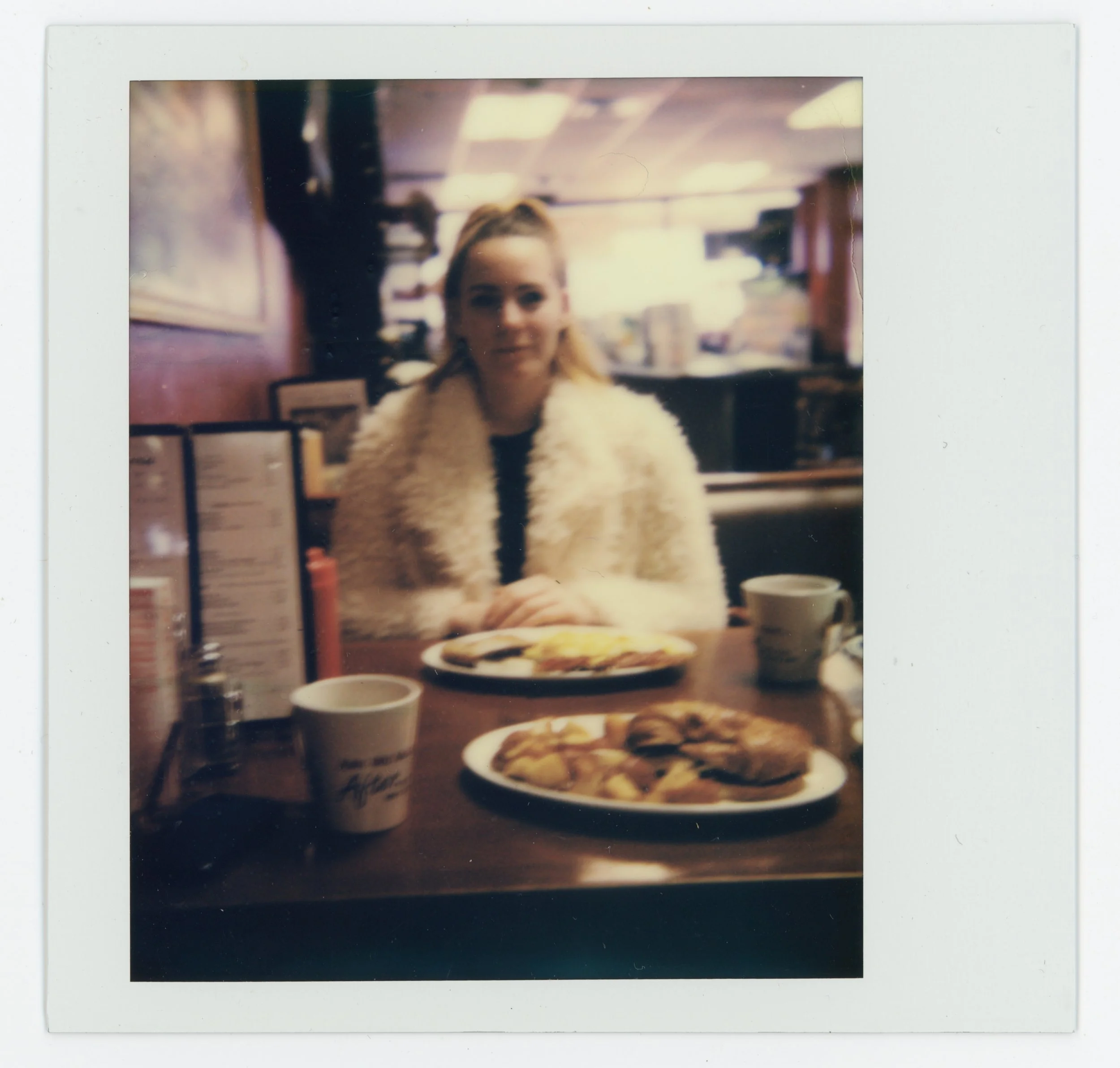 A woman sitting at a restaurant table with plates of breakfast food and cups, wearing a fluffy cream-colored coat, inside a cozy diner with a menu and warm lighting.