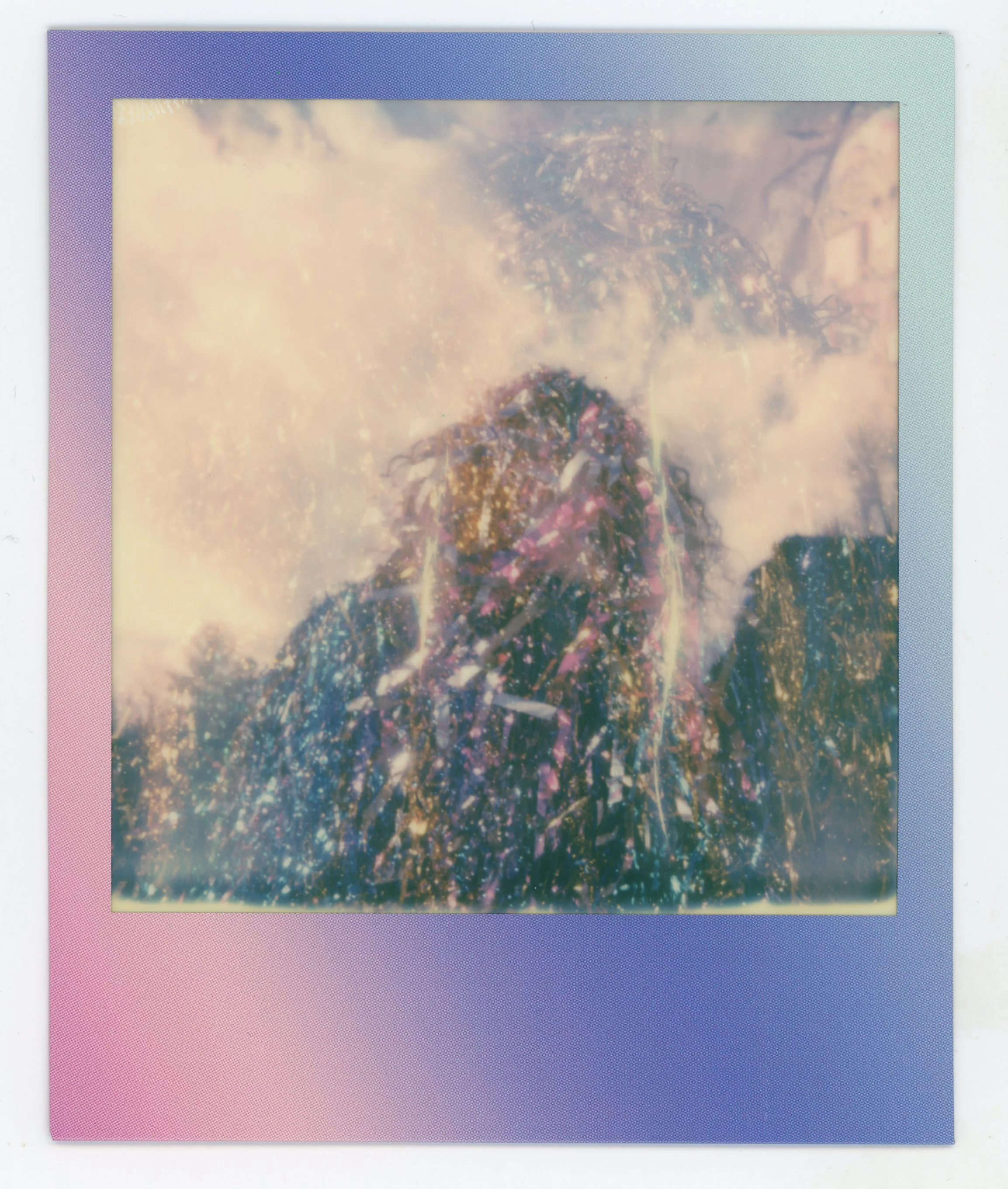 A polaroid picture with a gradient pastel-colored border of purple, pink, and blue. The photo inside is a dreamlike, blurry, and glittery representation of a person with long hair against a sky with clouds.