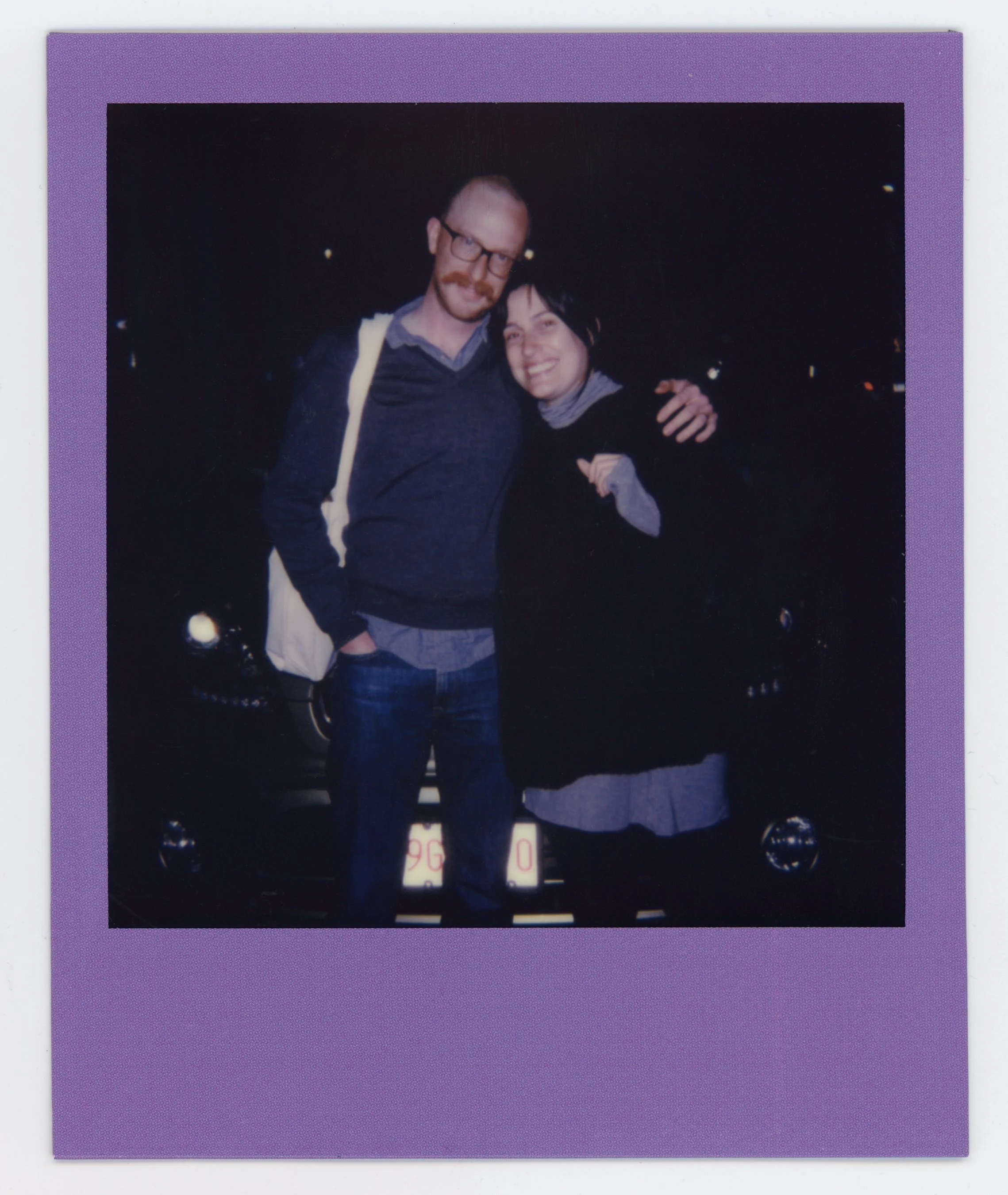 A man with glasses and a woman with black hair smiling and embracing outside at night, standing in front of a black car.