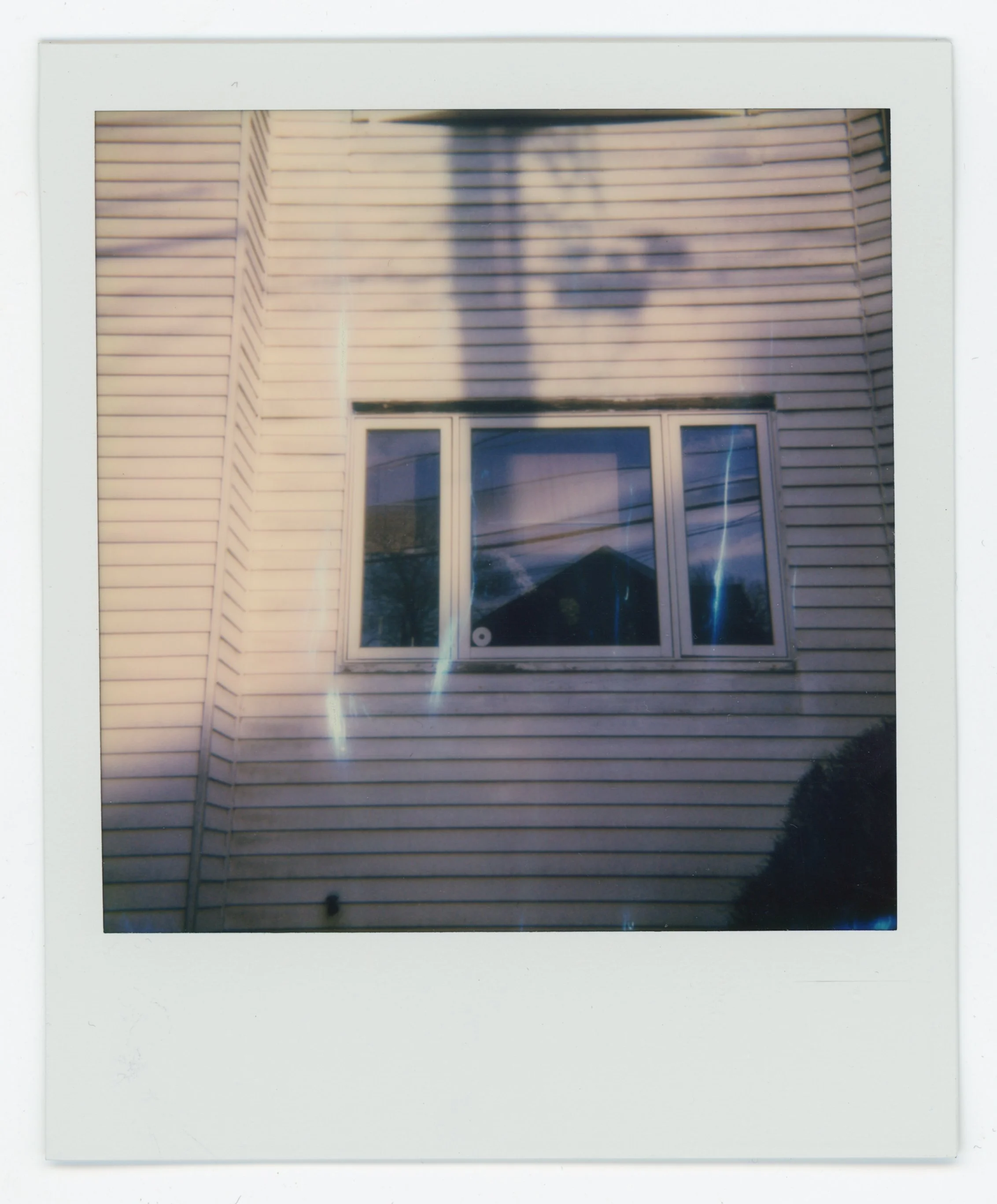 A window with three panes on a beige house with vinyl siding, reflecting the sky and a roofline, with shadows cast on the siding.