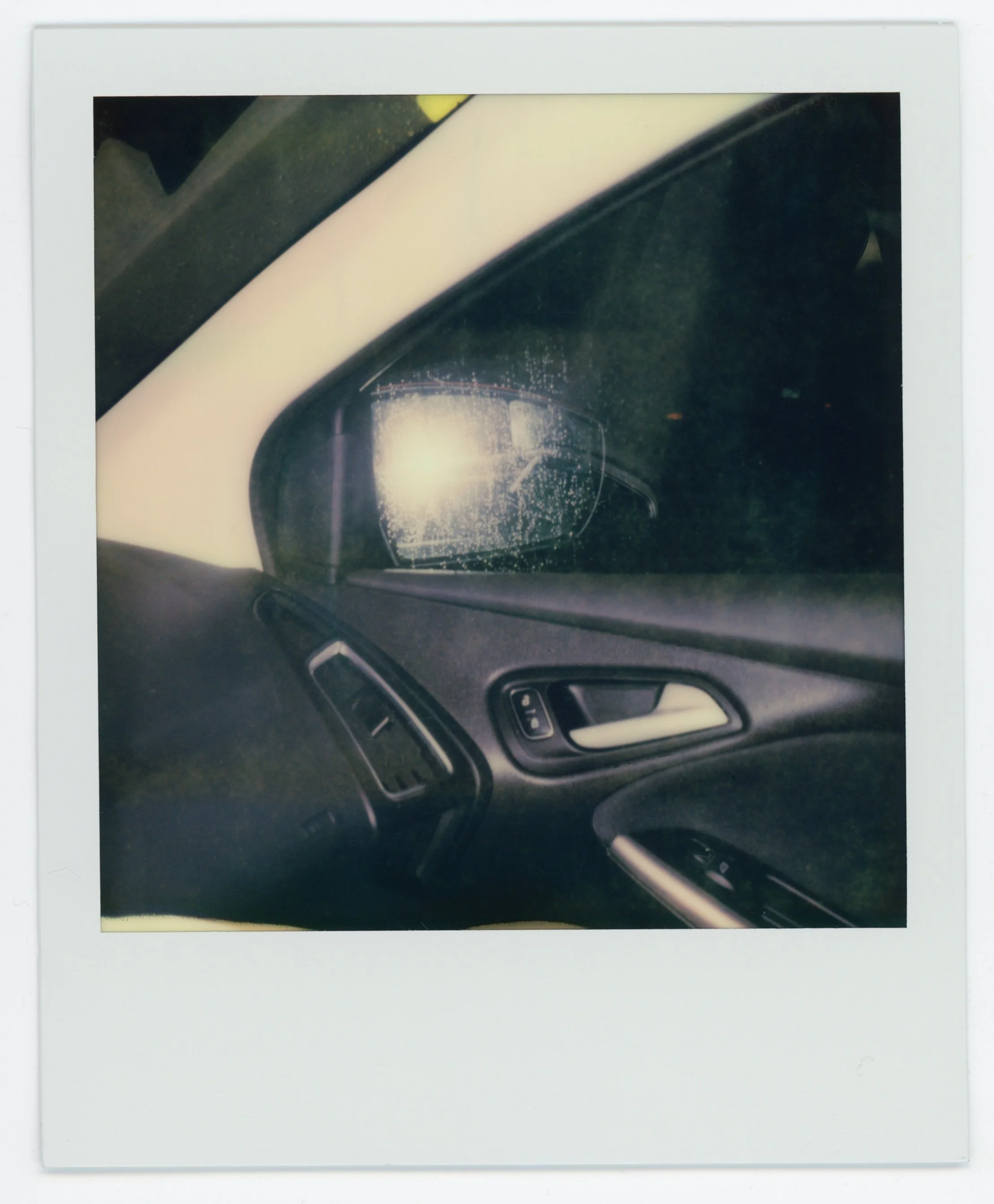 Polaroid photo of a car interior showing the driver's side door, window, and side mirror with raindrops on the window, taken at night.