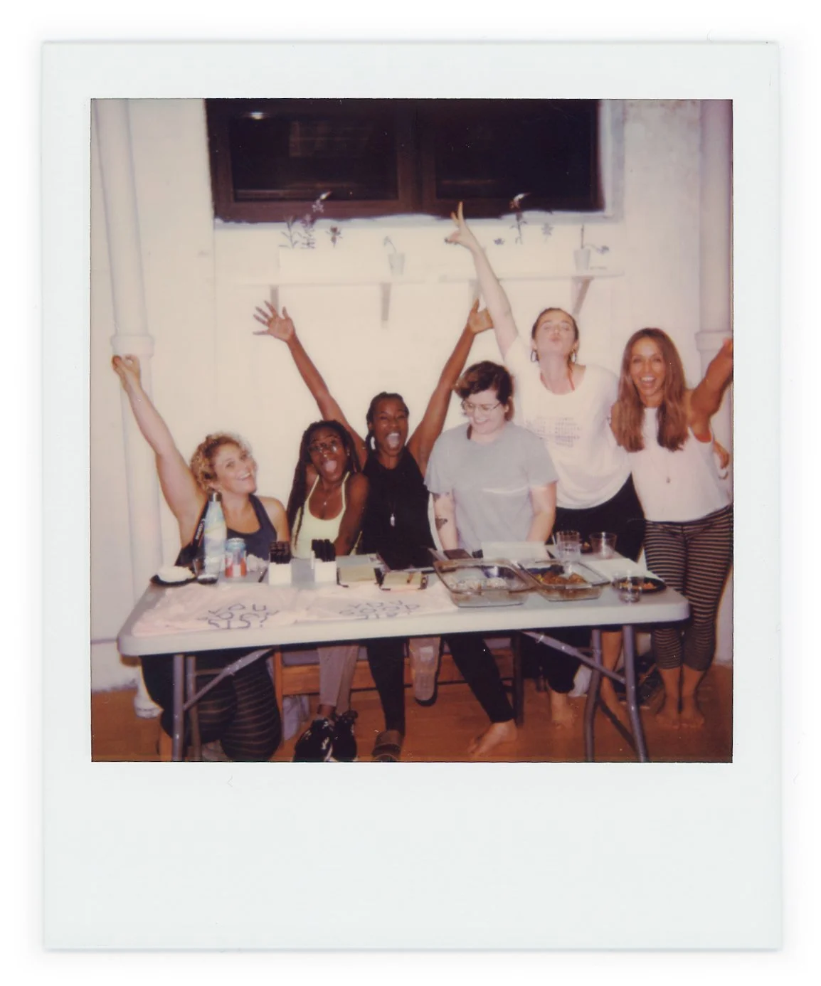 Group of six women celebrating around a table with food and drinks, some with arms raised in excitement.