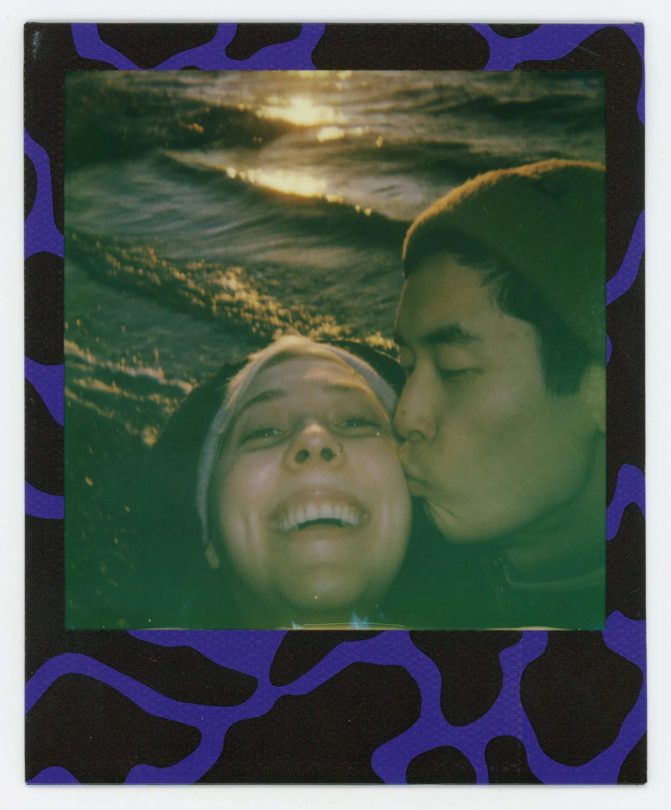 A Polaroid photo of two people, one kissing the other's cheek, with a sunset over the ocean in the background, framed by a purple and black patterned border.