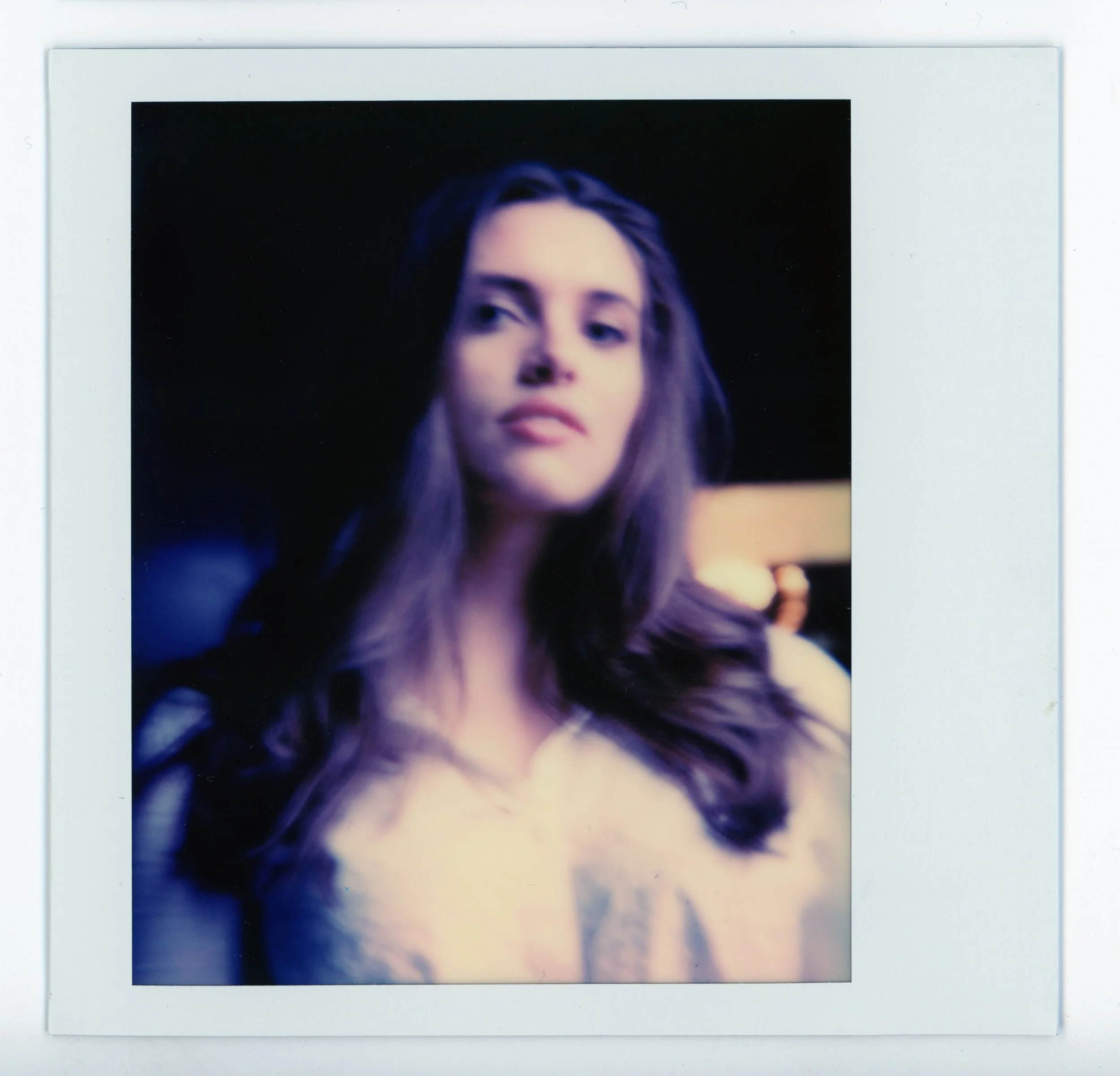 Polaroid photograph of a young woman with long brown hair, looking slightly to her right with a neutral expression, wearing a light-colored top, against a dark background.