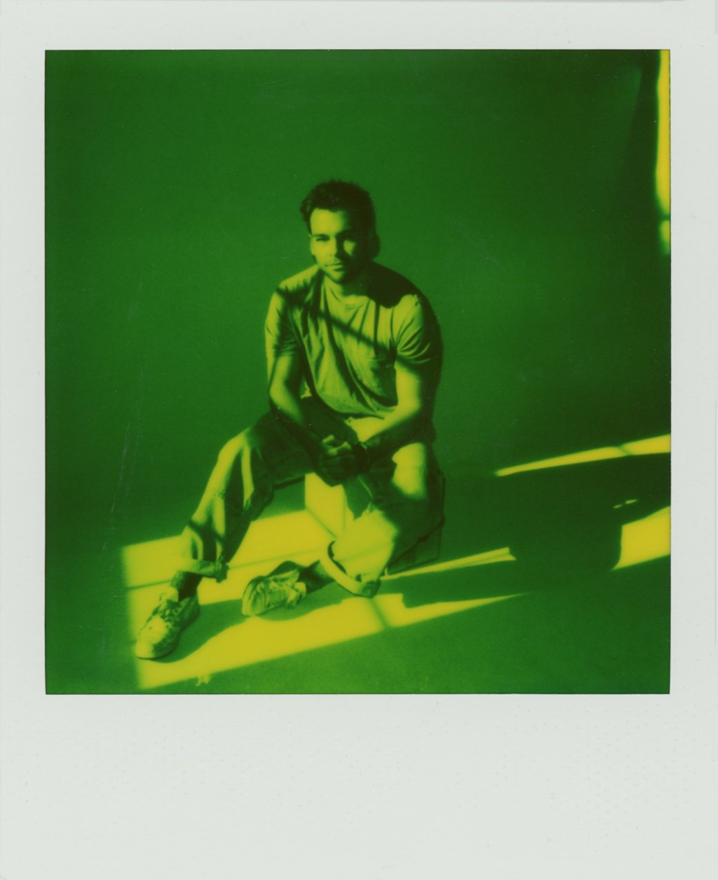 A young person sitting on a bench in front of a green wall, illuminated by sunlight.