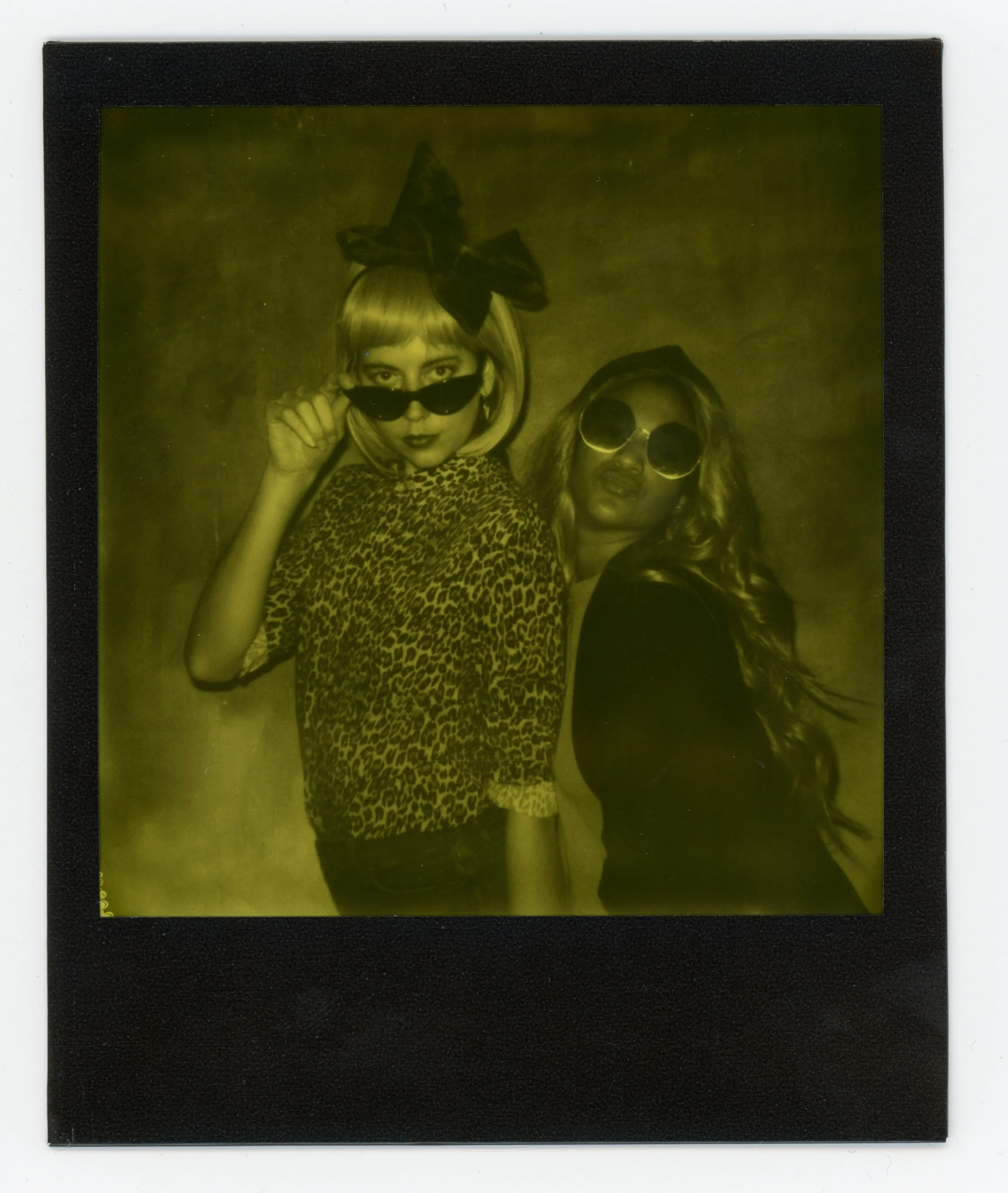 Two young women posing together, one wearing leopard print top and bow headband, the other with long wavy hair and round sunglasses.