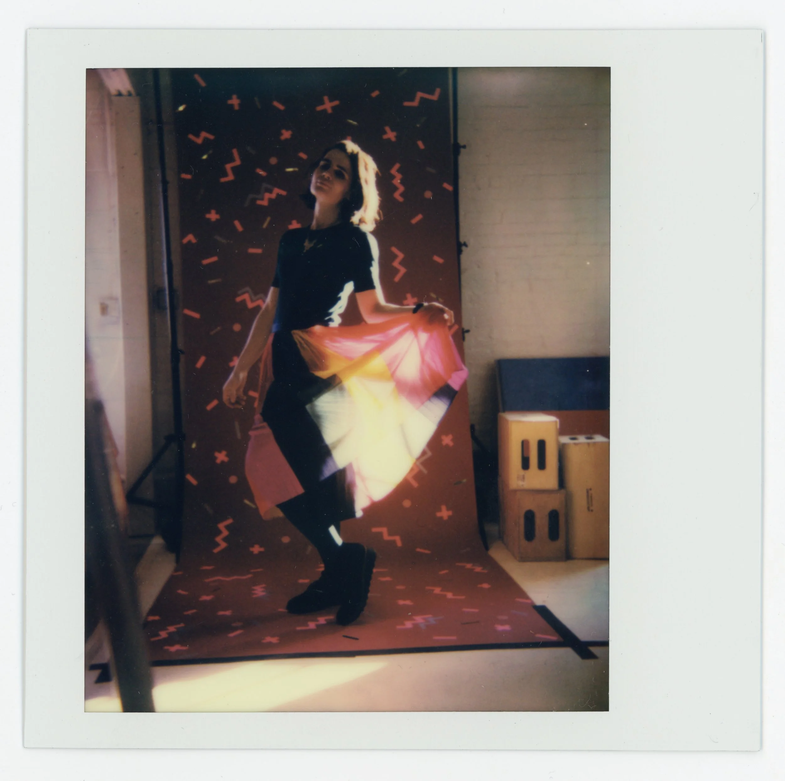 A woman posing in front of a colorful, abstract backdrop, holding a pink and yellow skirt, wearing black clothing and boots.