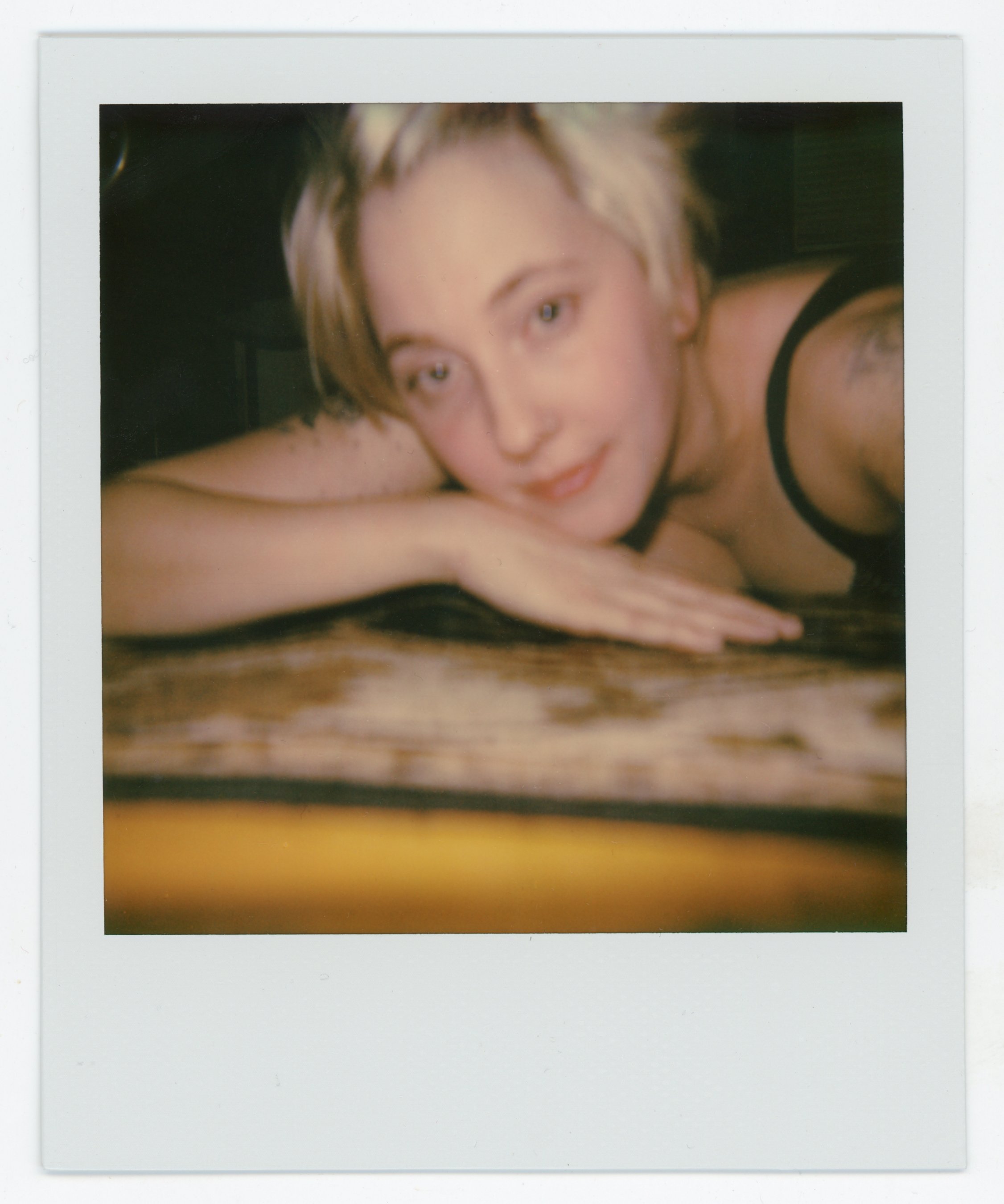 Polaroid photo of a woman with short blonde hair lying on a wooden surface, resting her head on her folded arm, wearing a black tank top, looking at the camera with a relaxed expression.