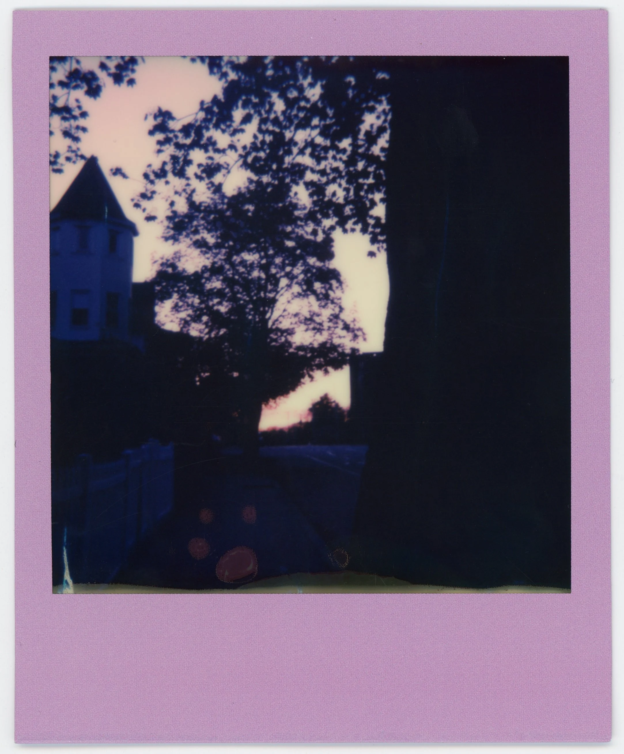 Silhouette of trees and houses at dusk or dawn with silhouette of a house with a pointed roof and window, and a fence along the side of the street.