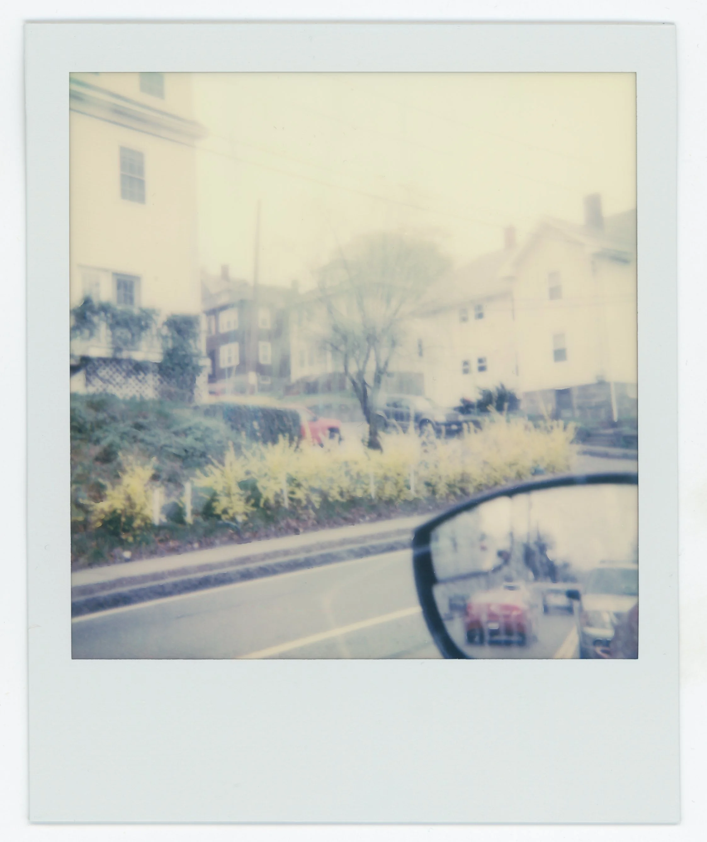 A blurry Polaroid photograph of a street scene with residential houses, parked cars, a tree, and a side mirror reflecting parts of the street.