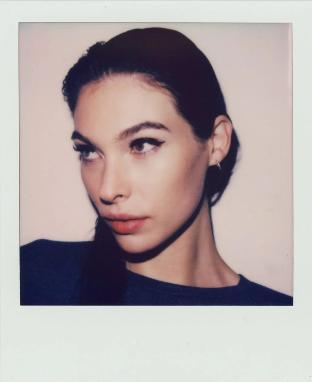 Close-up Polaroid photo of a woman with dark hair, winged eyeliner, full lips, and earrings, against a light background.