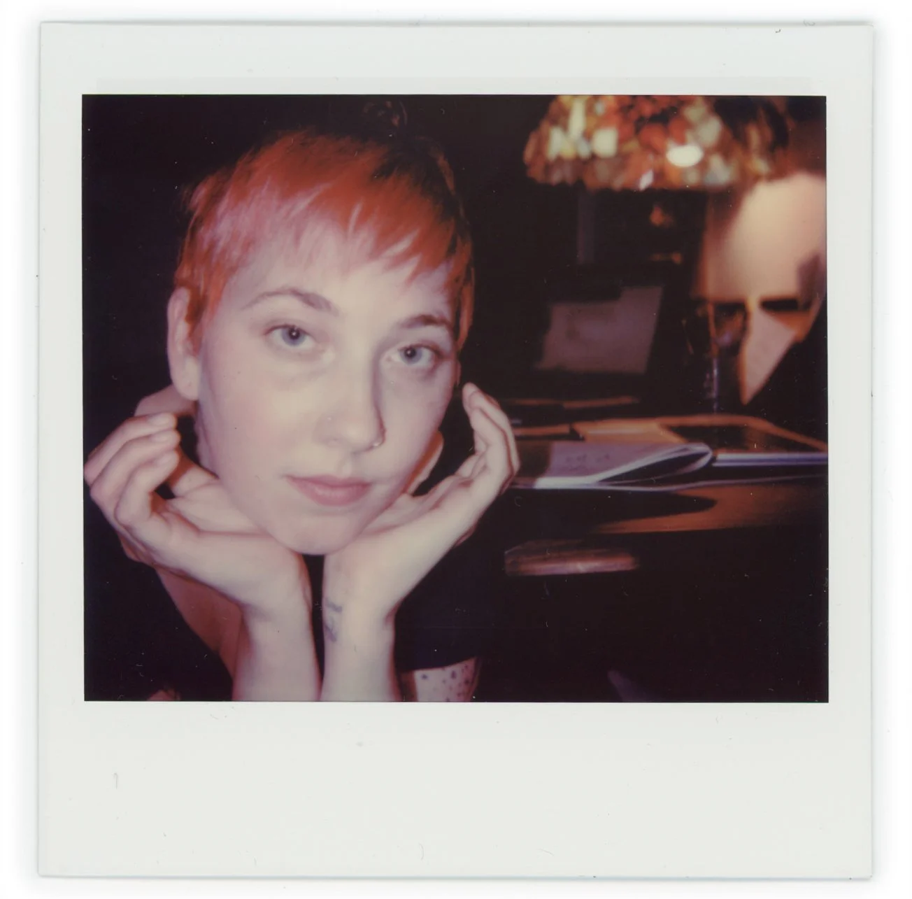 A young woman with short reddish hair resting her chin on her hands, sitting at a table with an open book, lamp, and a laptop in the background.