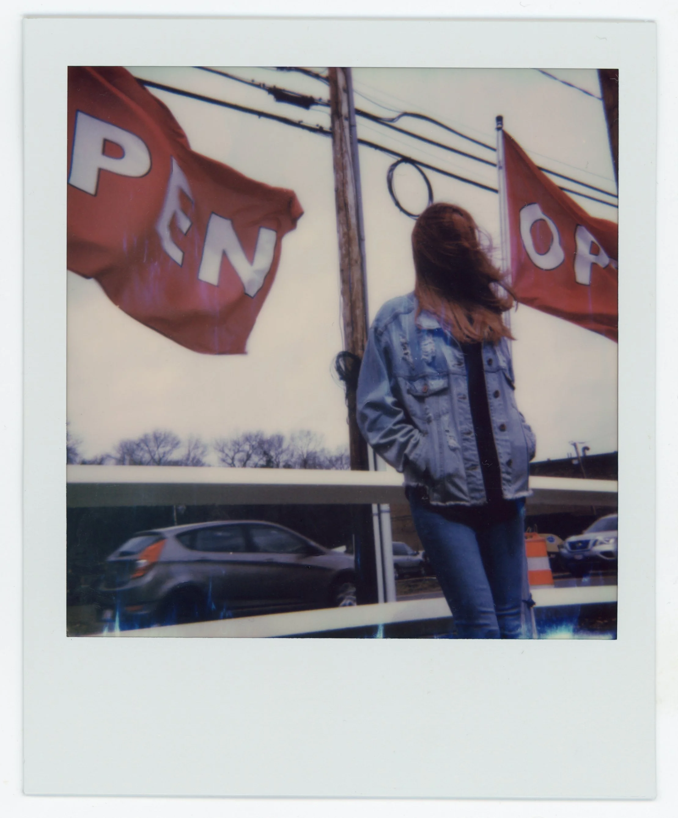A woman with long hair wearing a denim jacket standing outdoors near power lines and two red flags with 'OPEN' written on them, behind a reflective surface with cars passing by.