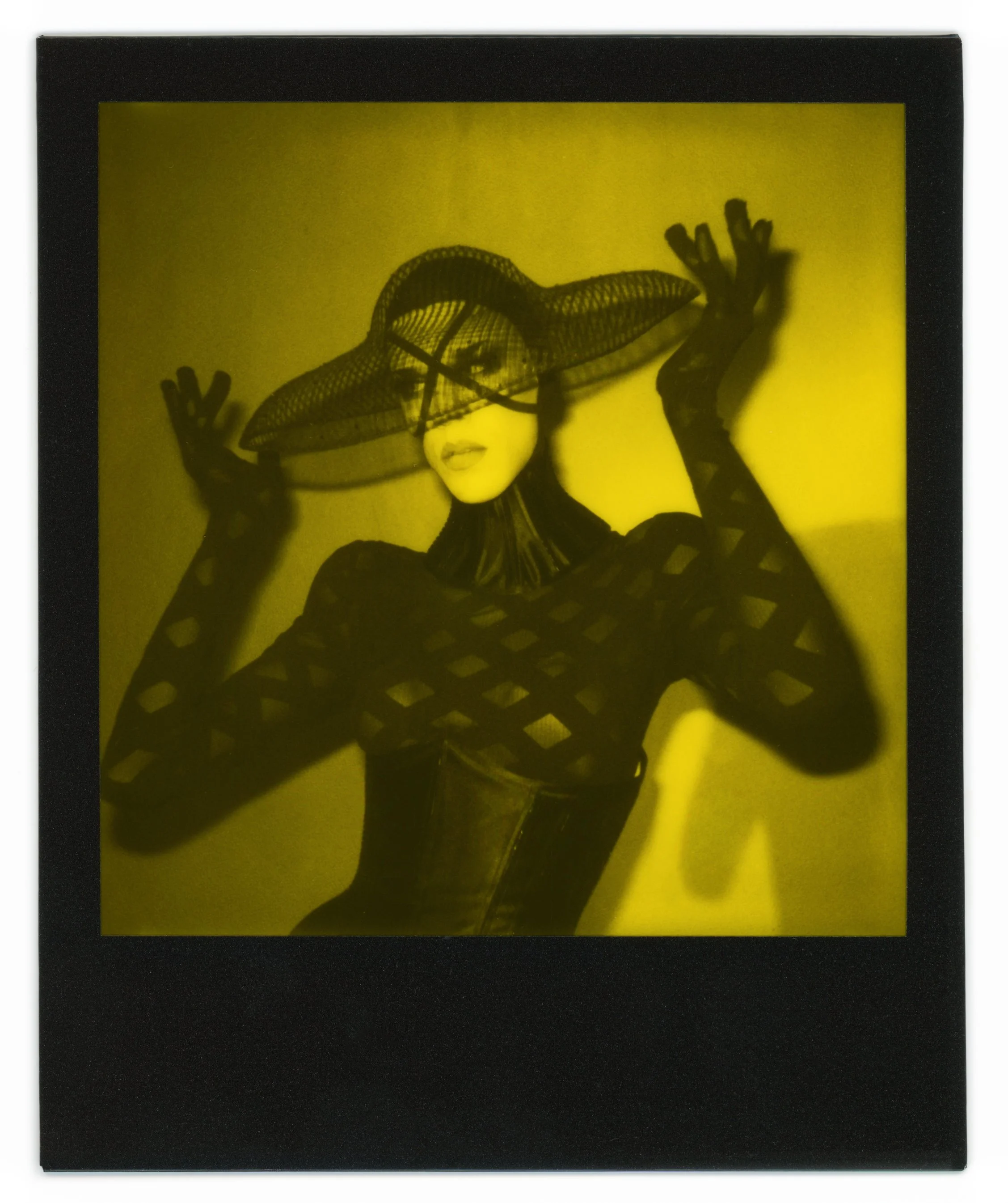 A woman wearing a wide-brimmed hat and a black fishnet top, posing with her hands raised near her head, with shadows cast on a yellow background.
