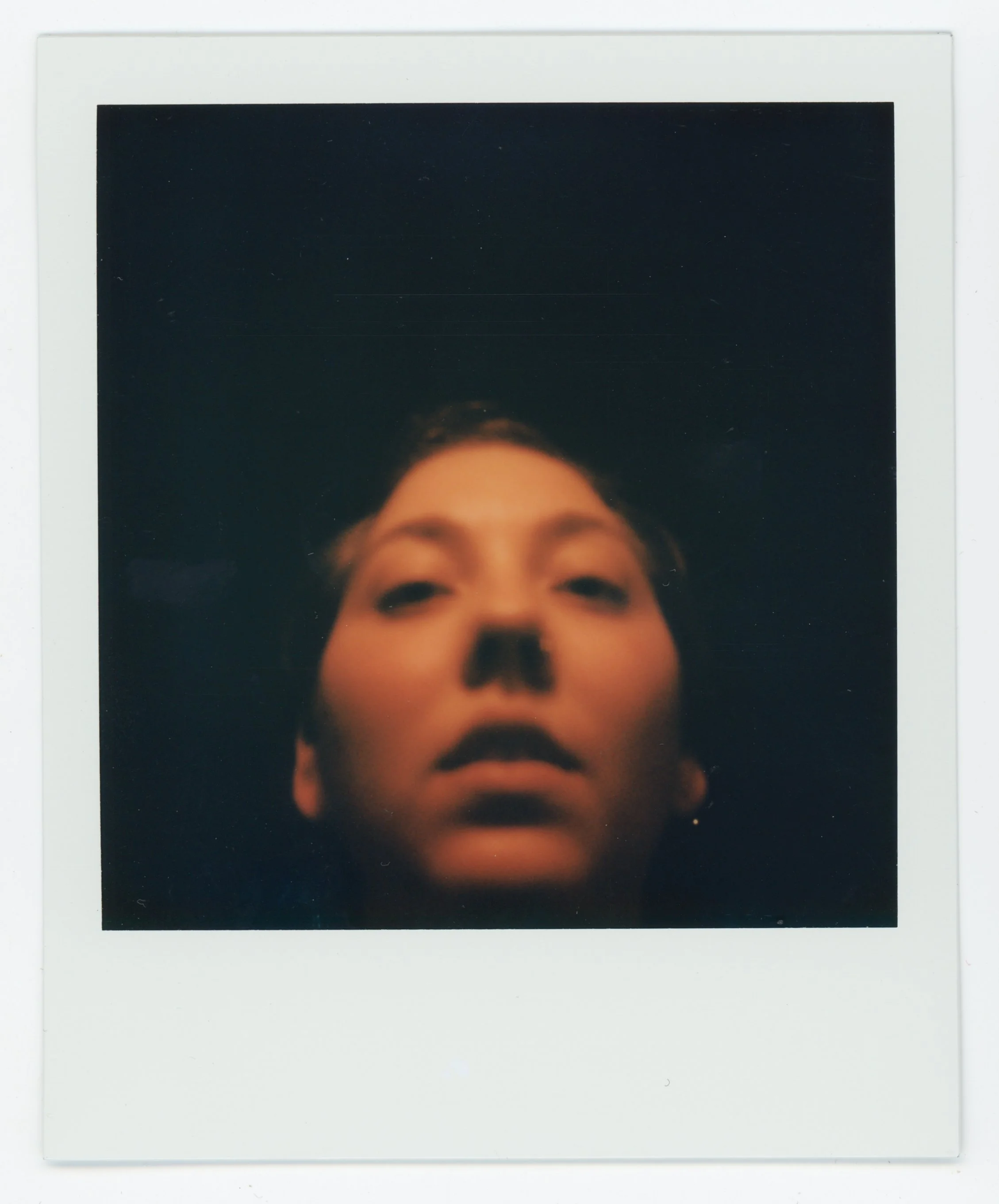 A close-up of a person's face with a dark background, with their nose painted black, resembling a skull, in an artistic or theatrical style.