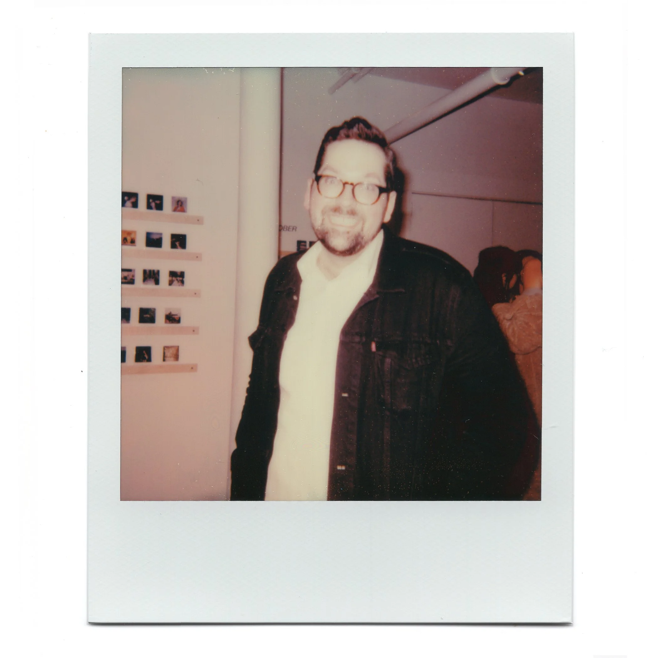 A man with glasses, a beard, and a smile, wearing a white shirt and a black jacket, standing indoors near a wall with small photographs displayed on it.