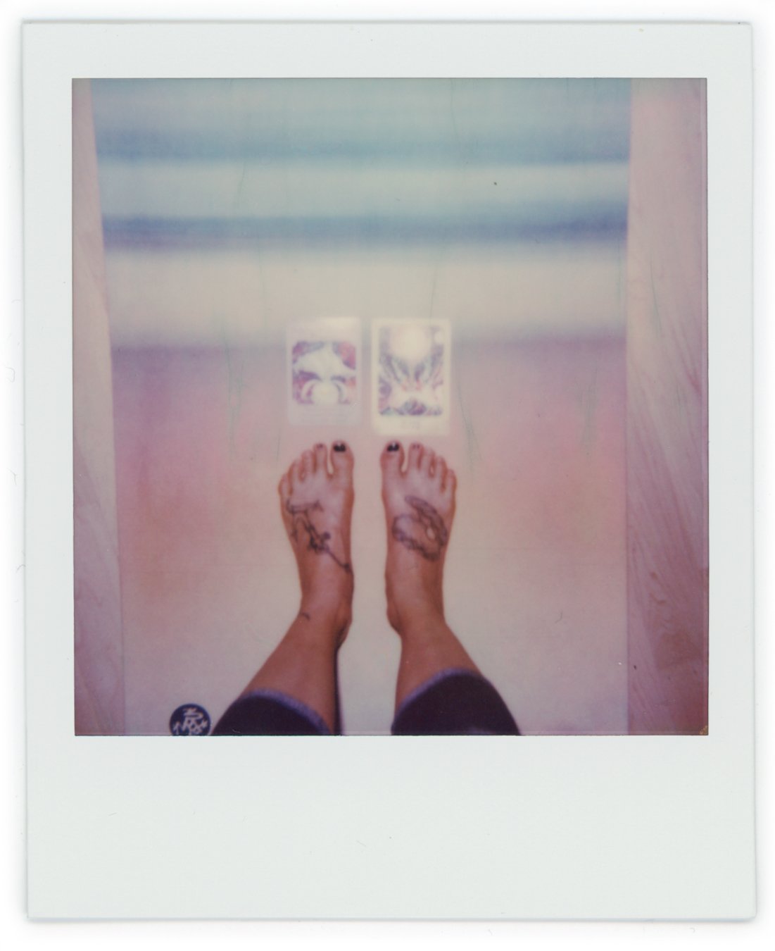 Polaroid photo showing a pair of feet with tattoos on the toes and ankles, standing on a pastel-colored floor, with two tarot cards facing up in front of the feet.