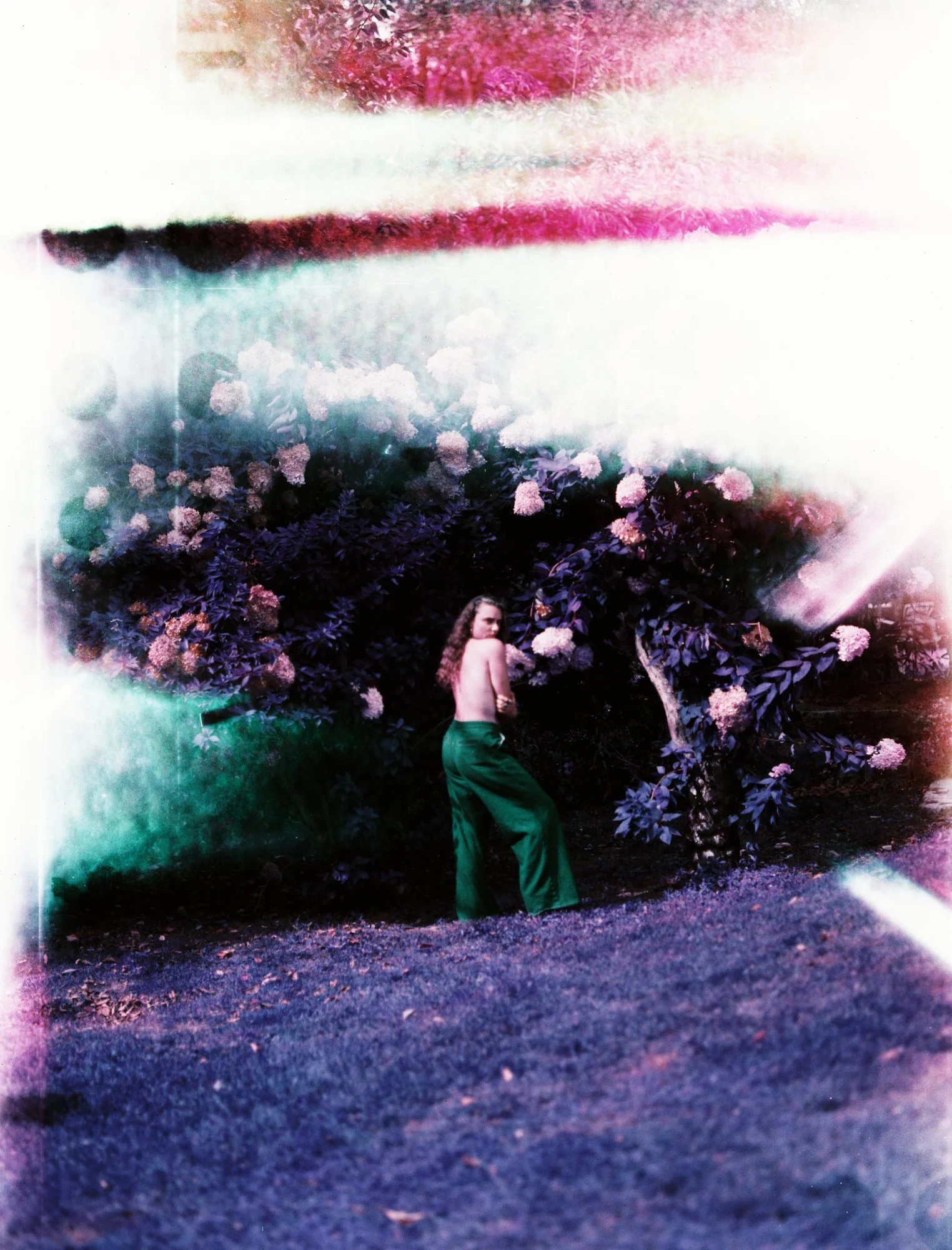 A woman with long, curly hair, wearing green pants and no top, standing outdoors in front of blooming pink and purple flowers, with a distorted, colorful effect.
