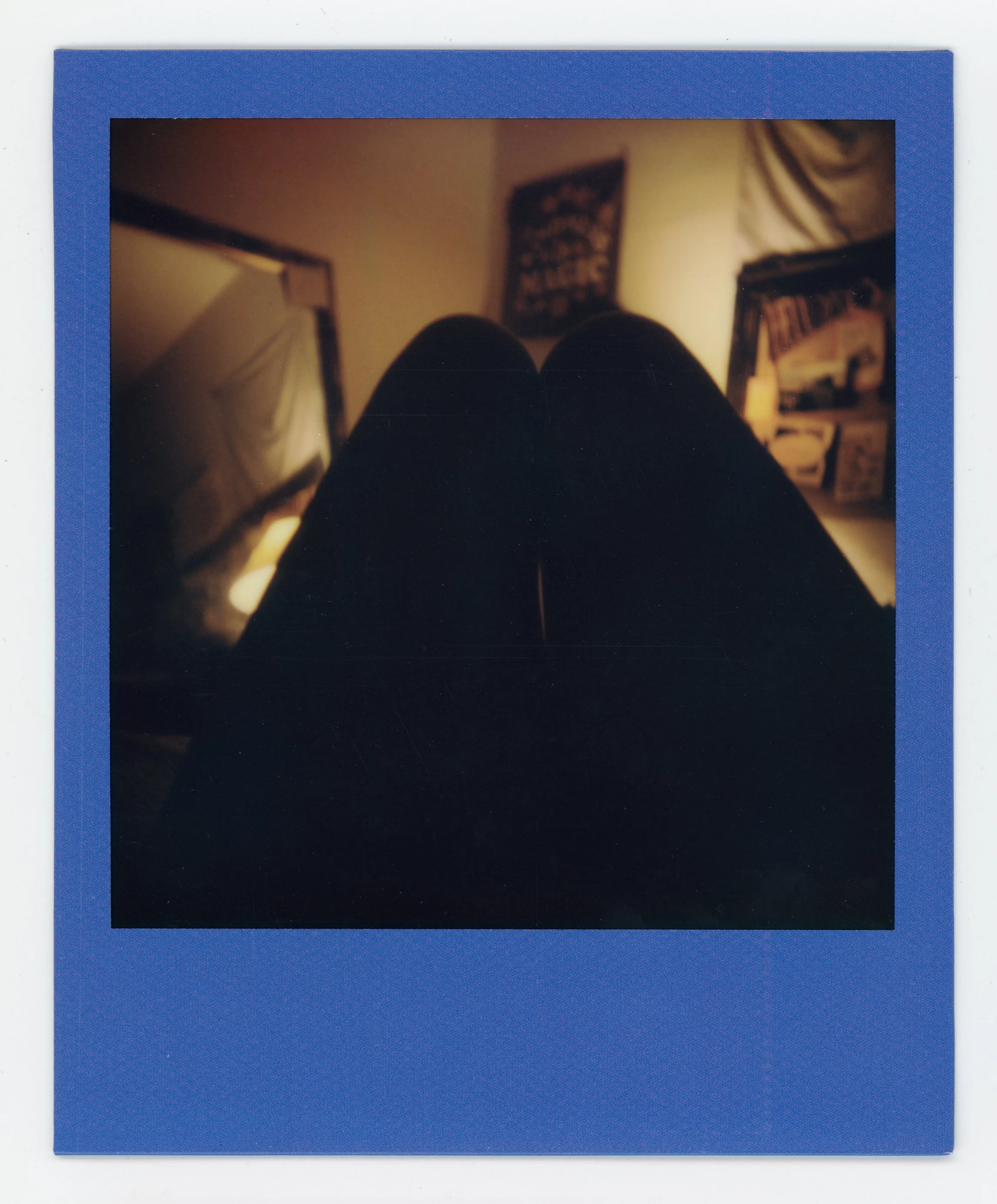 Polaroid photo showing a person's legs from a top view, with a bedroom background including framed pictures and posters.