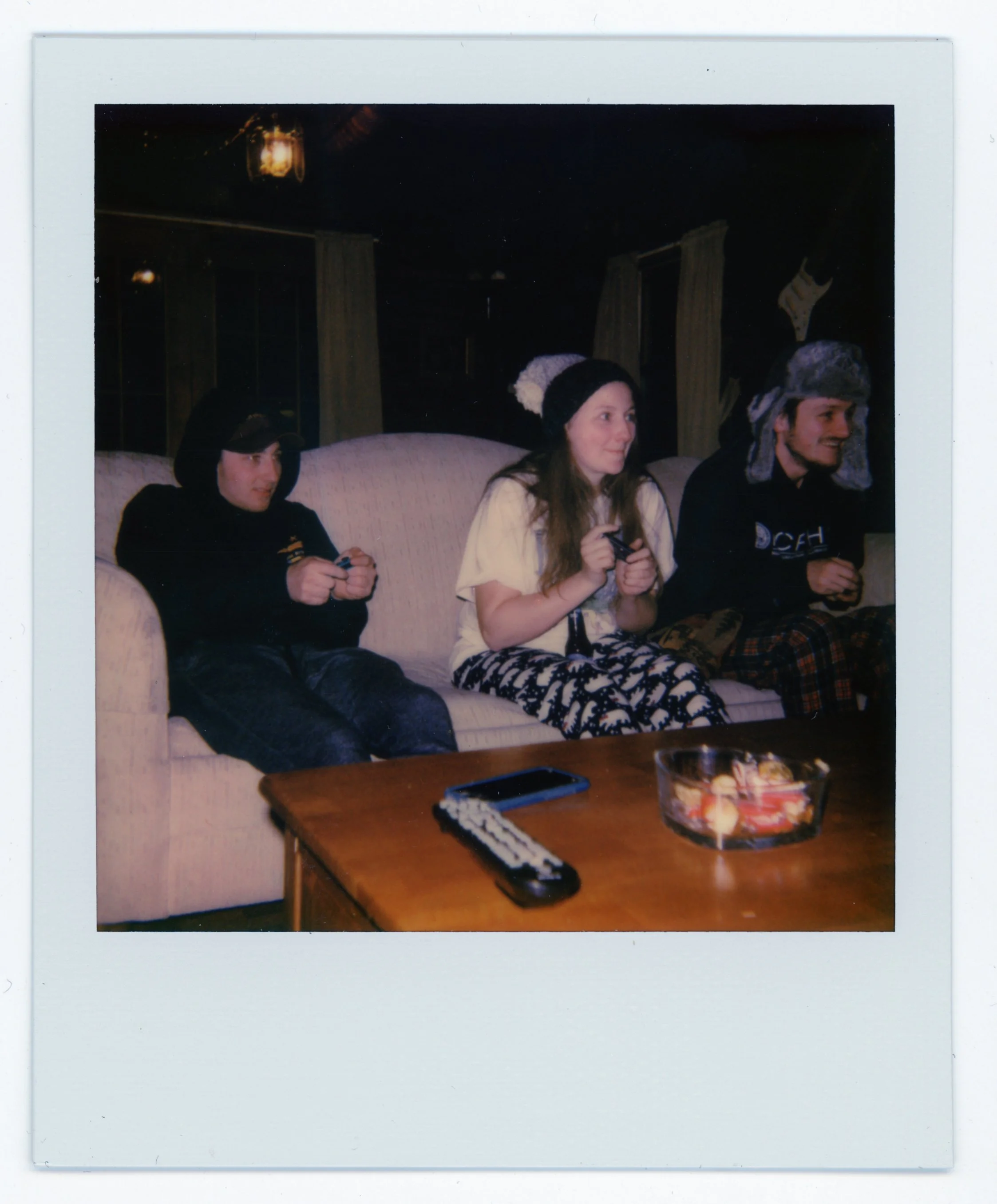 Three people sitting on a beige couch, wearing winter hats and pajamas, playing video games in a dimly lit room.