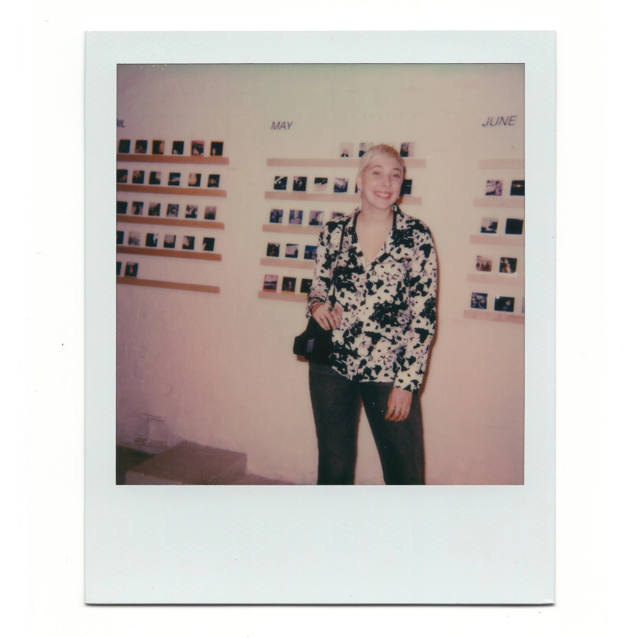 A woman with blonde hair smiling, standing in front of a wall with small photos arranged in rows under labels for months, including May and June.