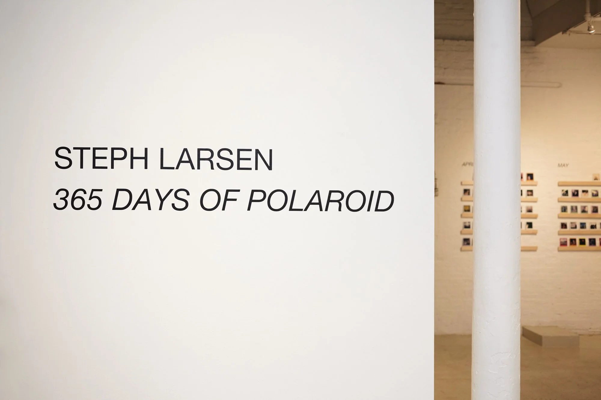 Exhibition sign reading 'Steph Larsen, 365 Days of Polaroid' inside a gallery with photos displayed on the walls.